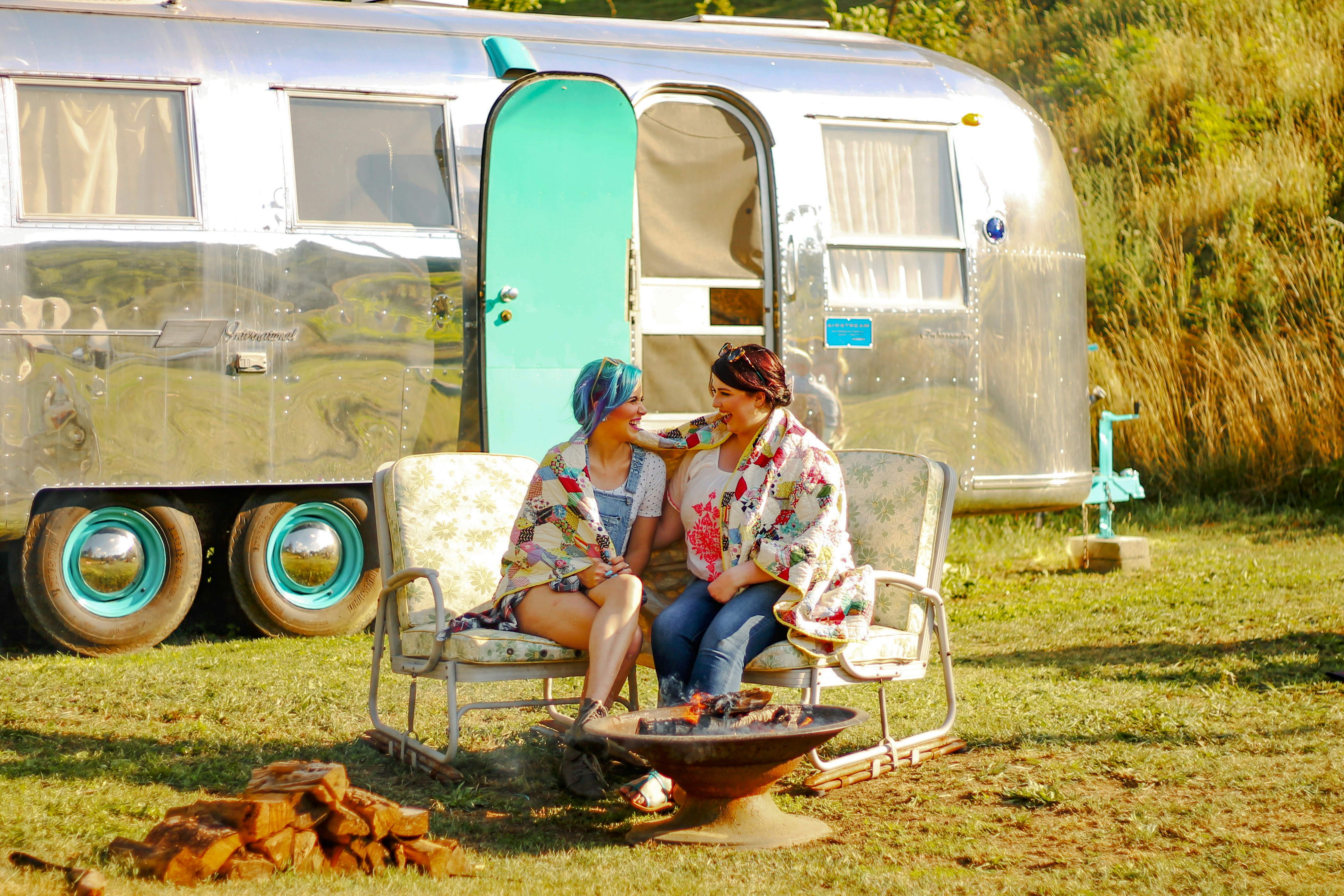 Two women laugh near a campfire while sharing a colorful blanket, with an Airstream trailer in the background