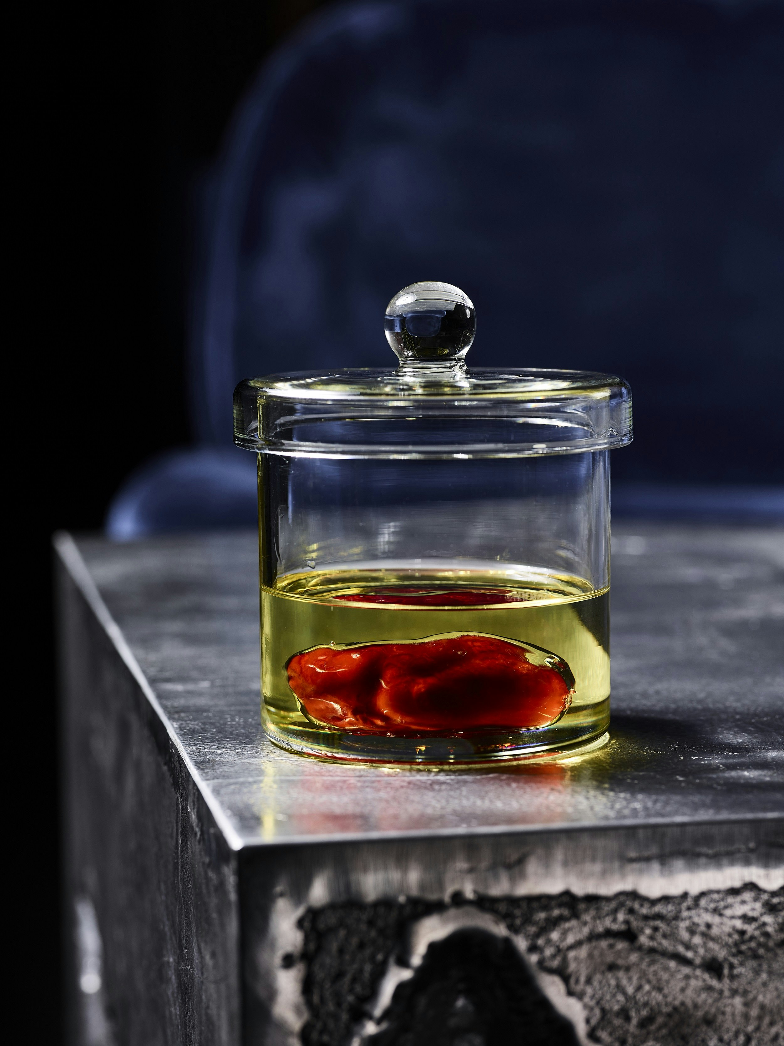 Sitting atop a dark marble table is a specimen jar filled with a golden liquid with a globular, red piece of food in it.