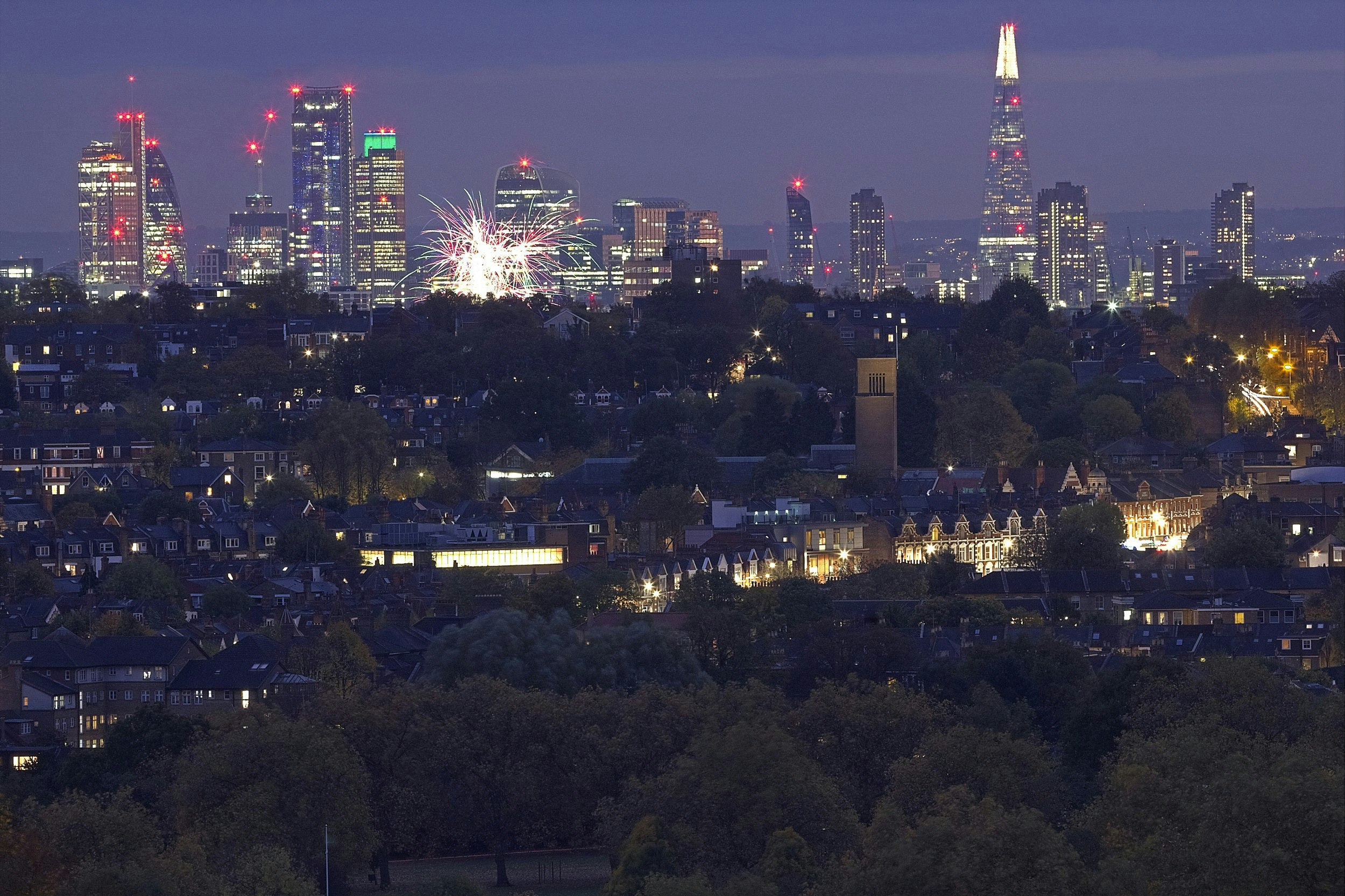The view of London from Alexandra Palace. Residential streets in the foreground give way to London's skyscrapers at dusk, with some fireworks visible in the sky