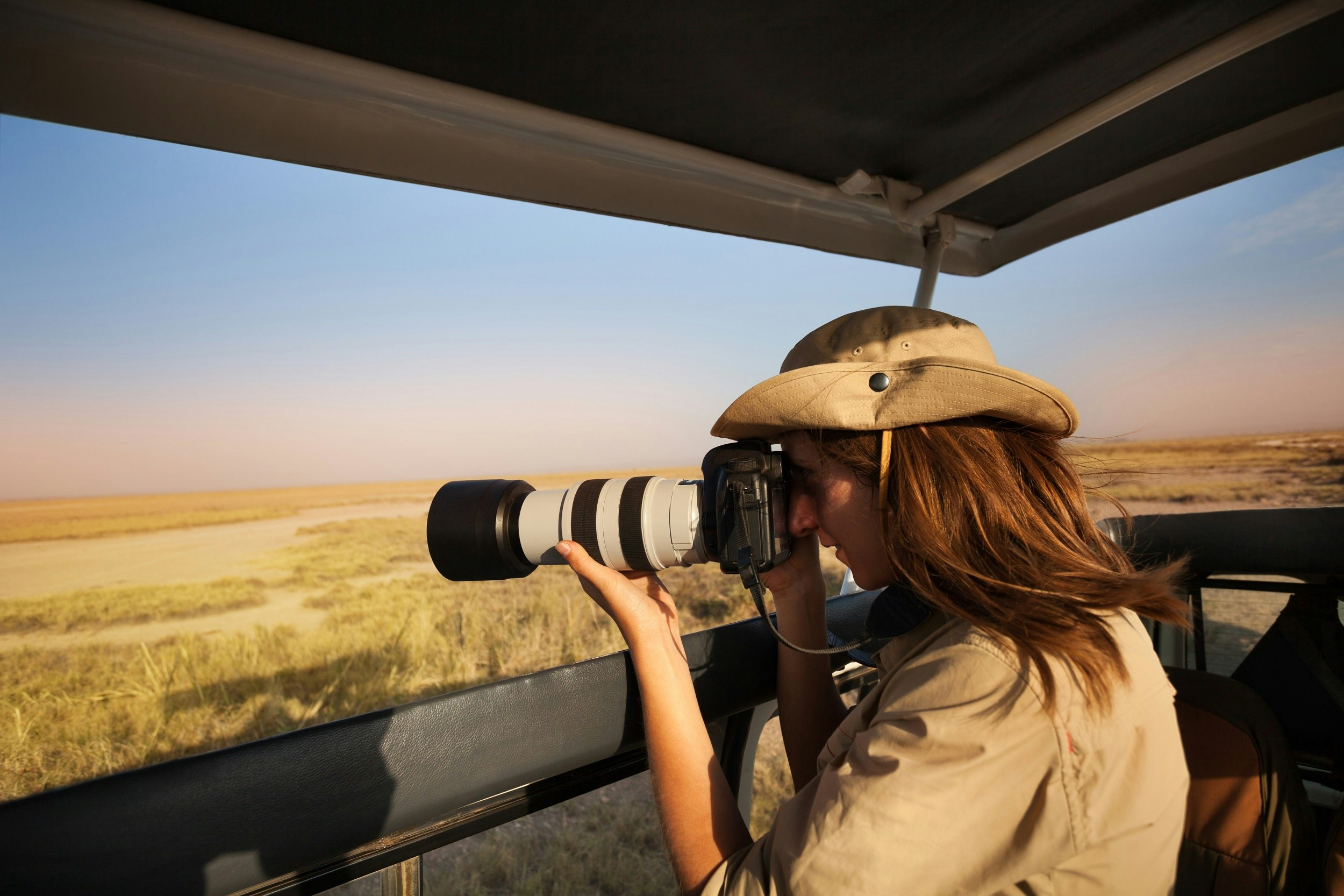 A woman tourist taking photo of savannah with a professional camera aboard safari jeep in Amboseli National Park; the truck has a pop-up roof, so she is standing in the jeep with the roof above her head.