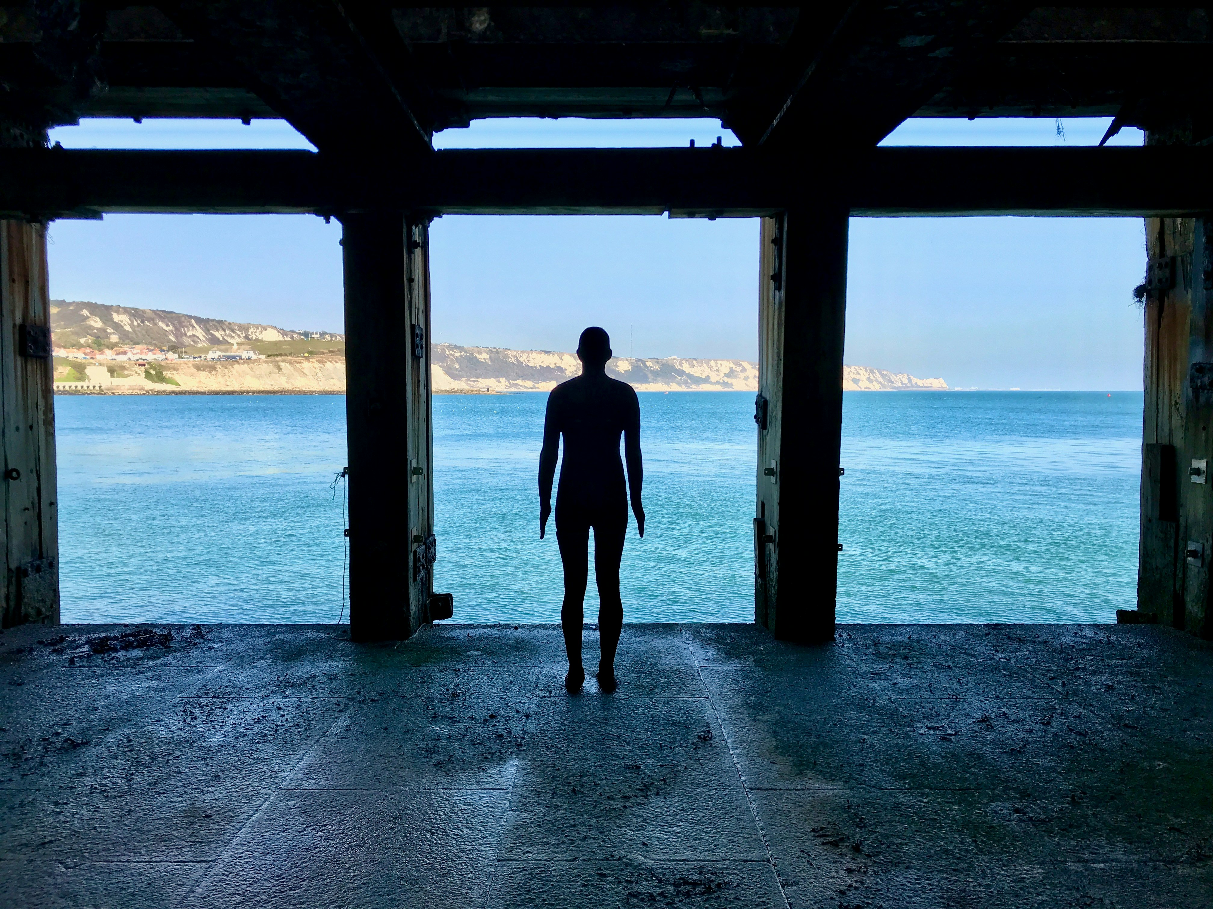 A cast iron figure by sculptor Antony Gormley stands in a shell of a building, looking out upon cliffs and the sea in Folkestone.