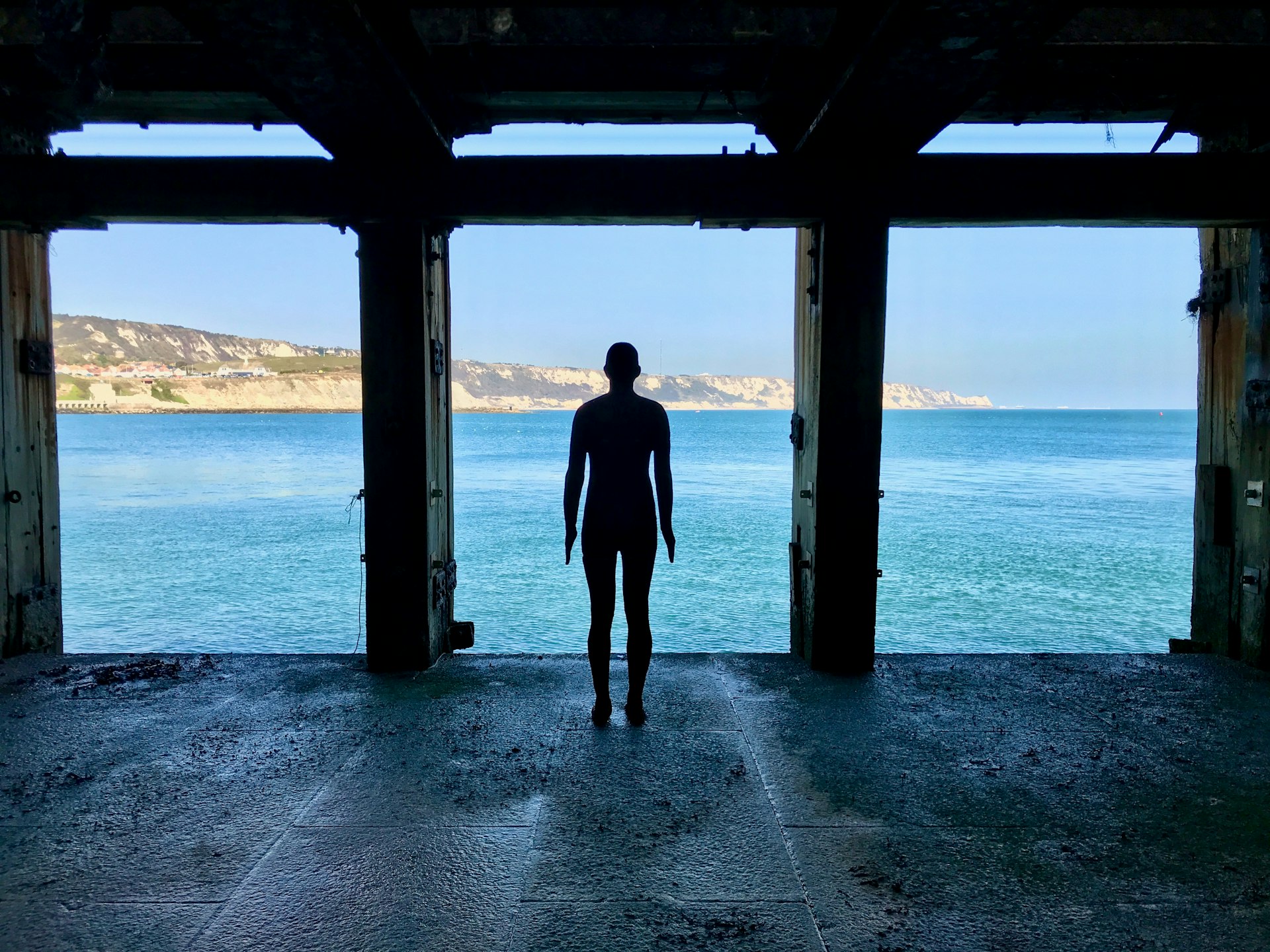 A cast iron figure by sculptor Antony Gormley stands in a shell of a building, looking out upon cliffs and the sea in Folkestone.