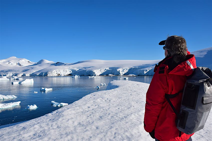 Michael Ballard stands with his back to the camera in a red cruise-line parka and gray dry-bag style backpack gazing out across the snow and ice fields of Antarctica 
