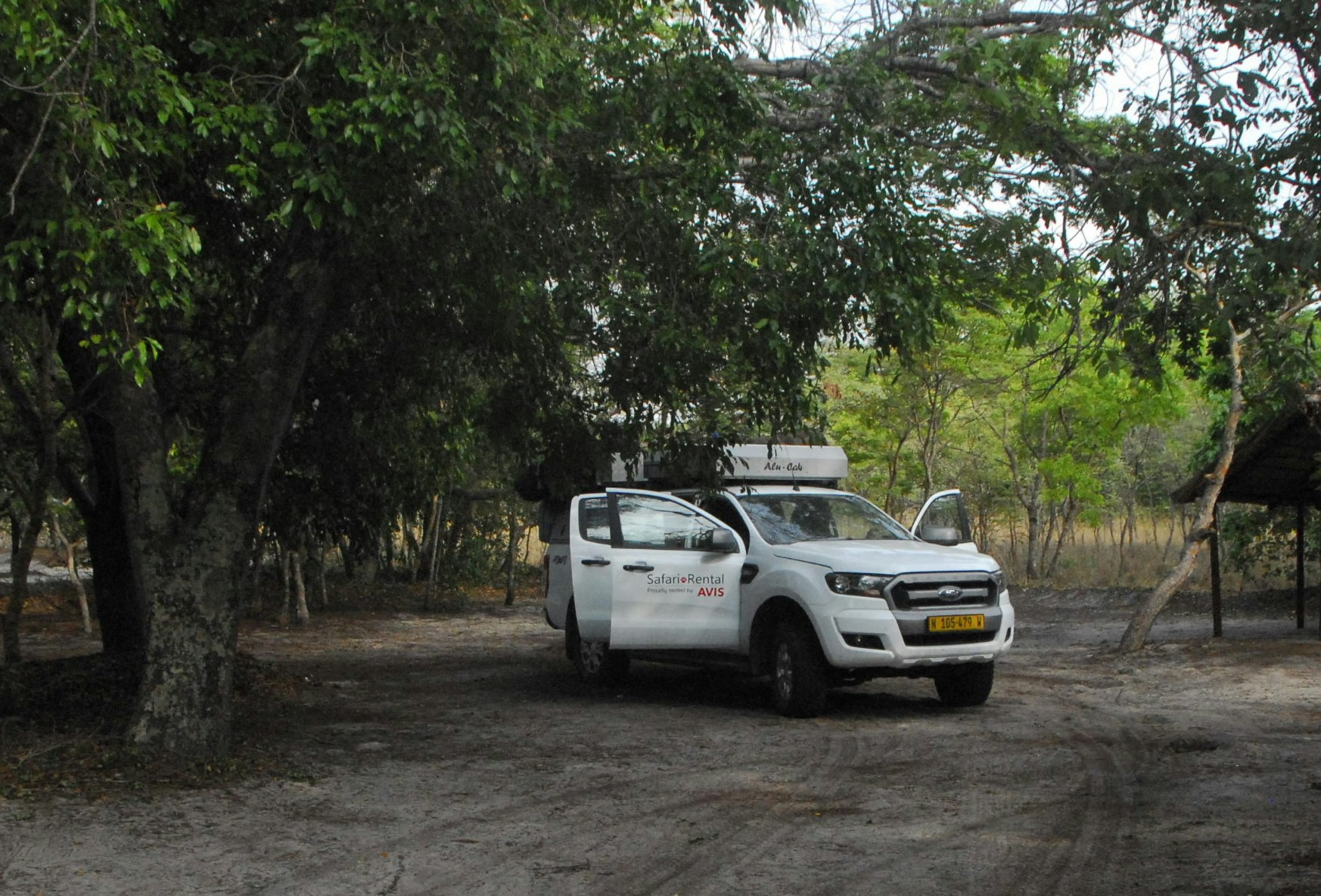 A white Ford truck, with its doors open, sits on a dirt path surrounded by trees and a small thatched structure; two fold-down tents are on the truck's roof.