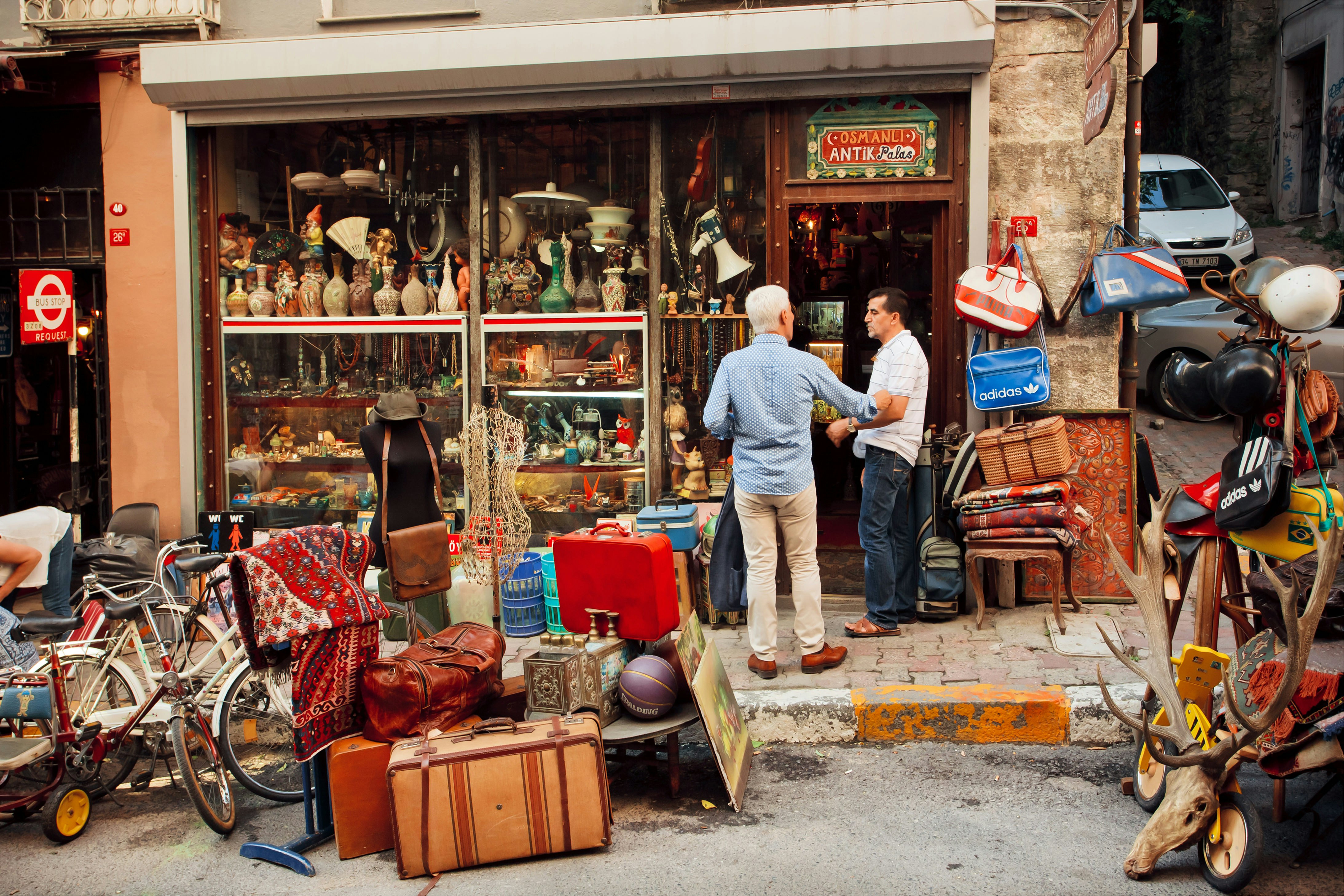 Two men are chatting in the doorway of an antiques shop in Çukurcuma, Istanbul. The pavement outside is piled high with various goods, from suitcases to bicycles. 