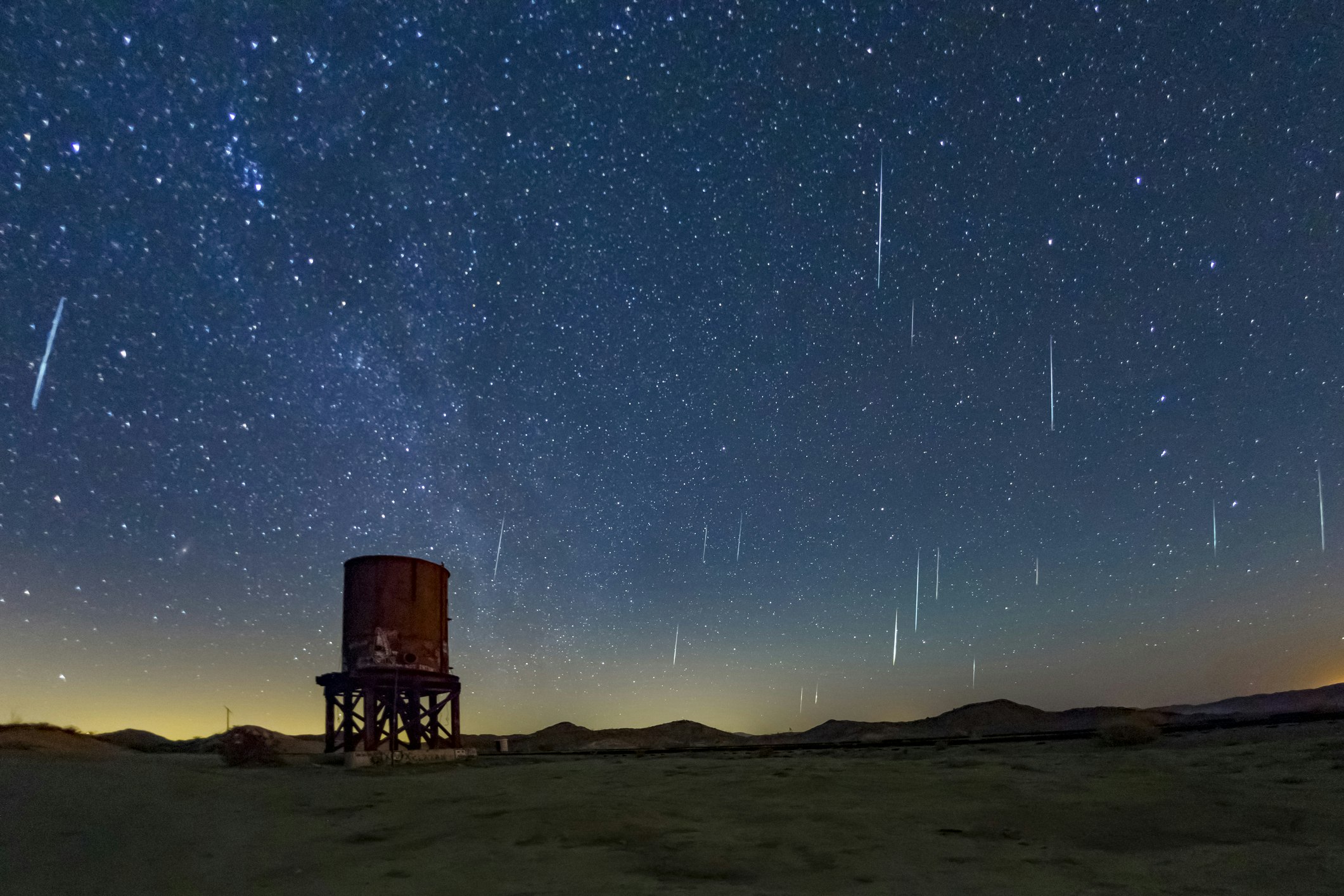 The Geminids Meteor Show in 2017 falls over Anza Borrego Desert State Park. The sky is a bright, warm indigo blue dotted with white and light blue stars and streaks from the falling meteors. The horizon is a light yellow or deep orange and framed by low mountains and hills. In the mid ground is a red water tower, part of the old Dos Cabezas Siding 