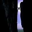 Silhouette of man rock against a purple sky; he is wedged between two vertical cliff faces that are also silhouetted.