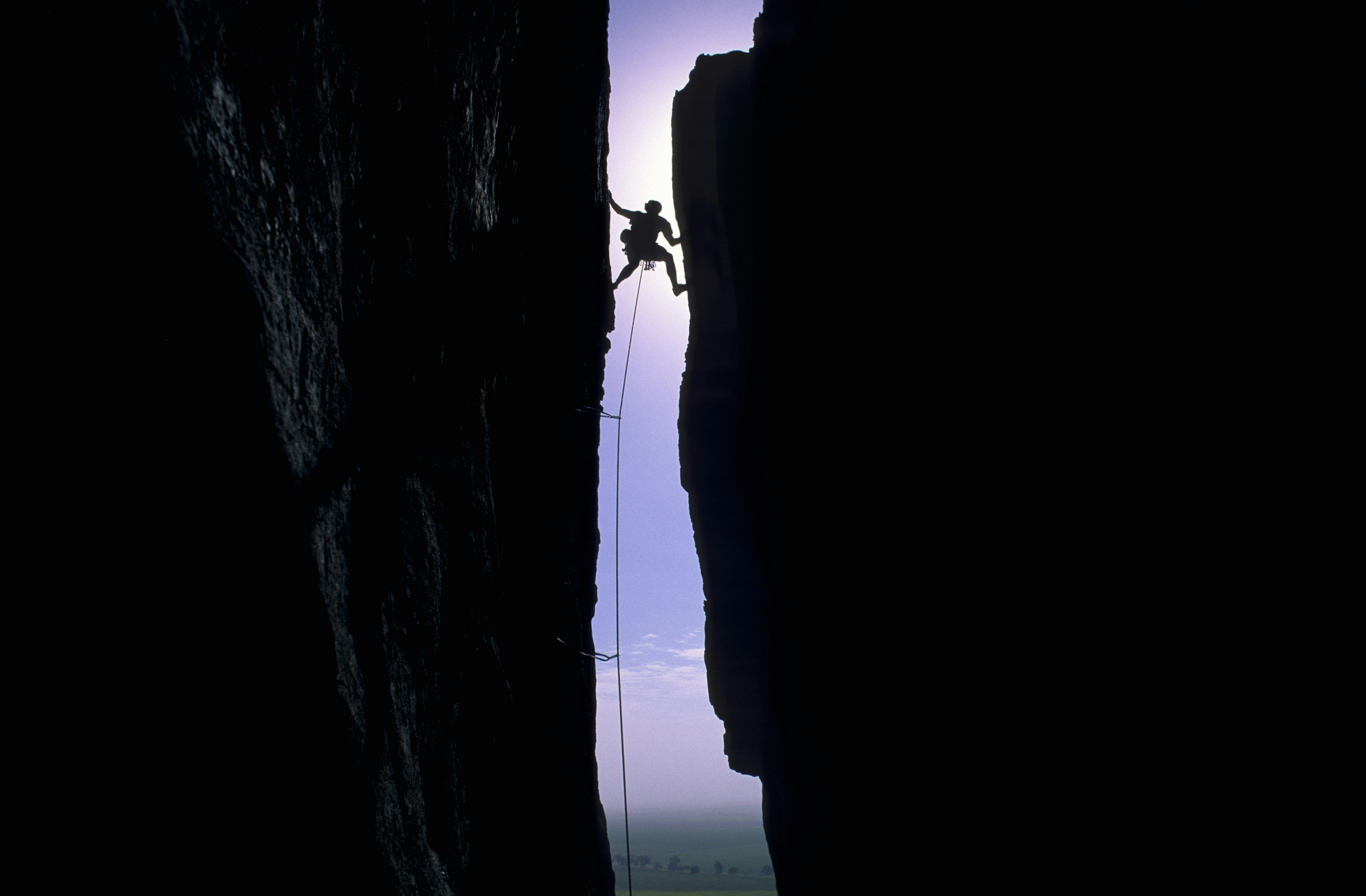 Silhouette of man rock against a purple sky; he is wedged between two vertical cliff faces that are also silhouetted.