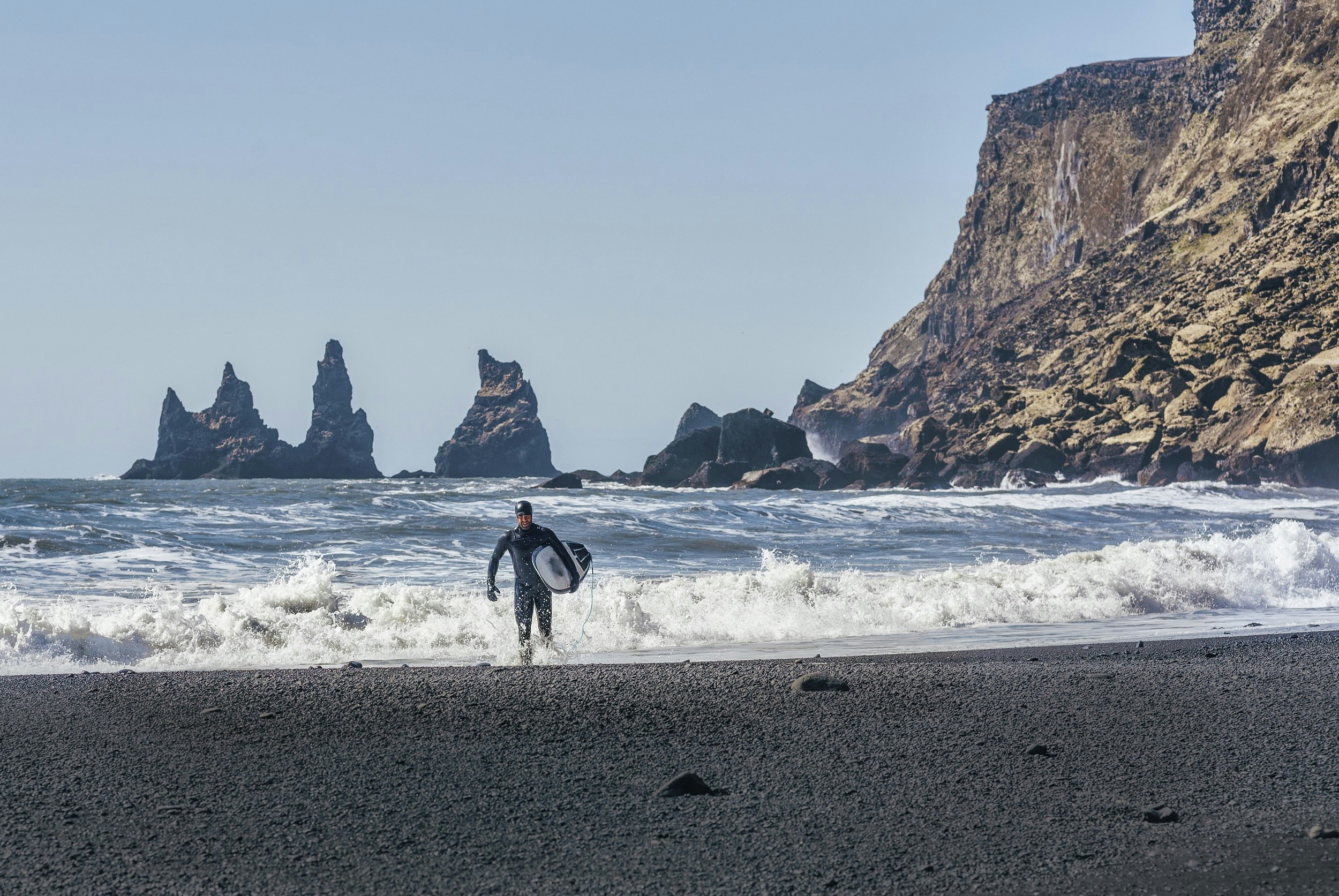 A surfer walks out of the surf and onto a black beach with his board in hand; in the background are several jagged islands bursting from the sea.