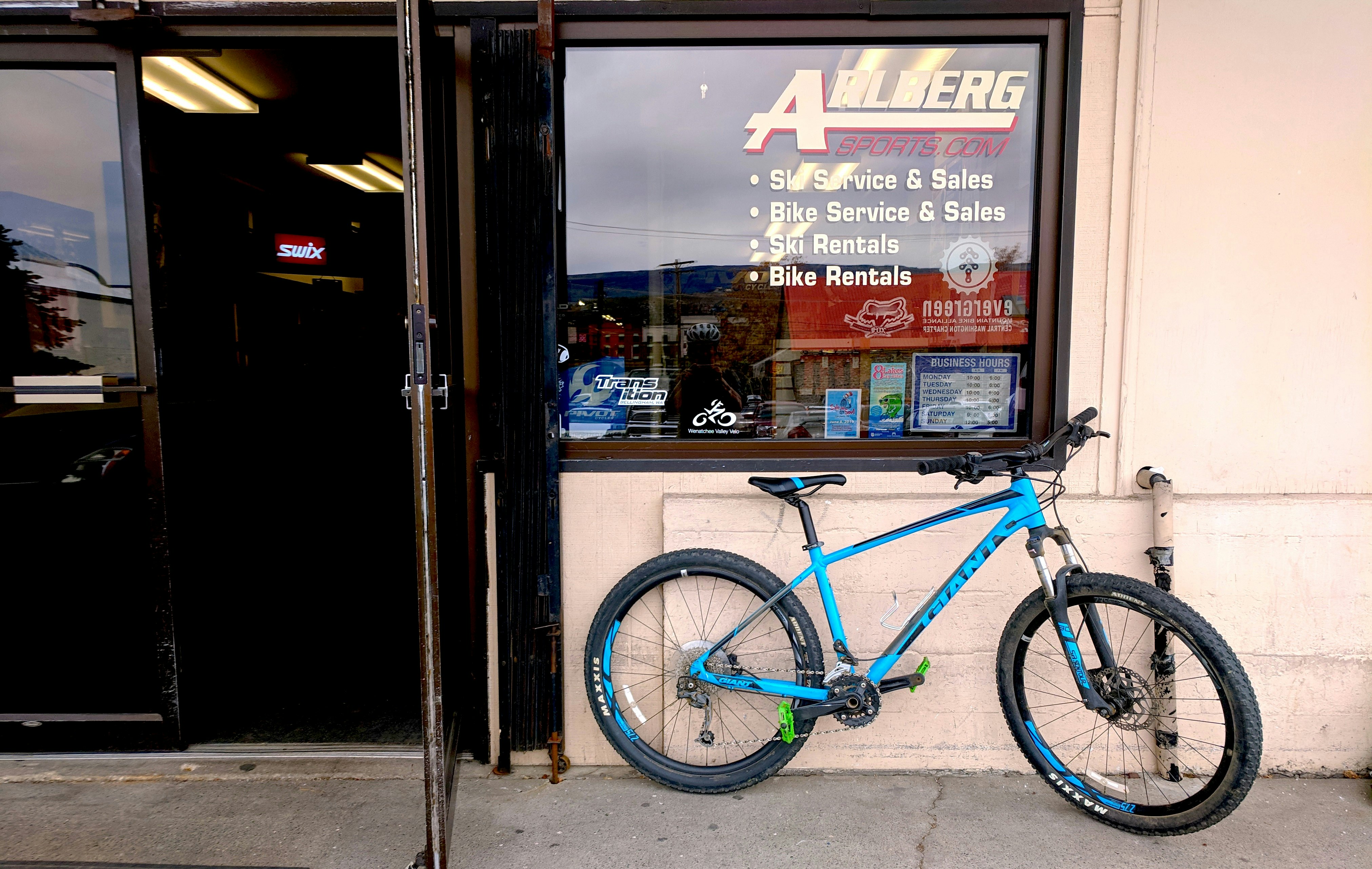 A blue mountain bike sits outside the entrance to Arlberg Sports in Wenatchee, Washington, the name of which is painted in cream letters on a window over the bike along with information about ski and bike rentals