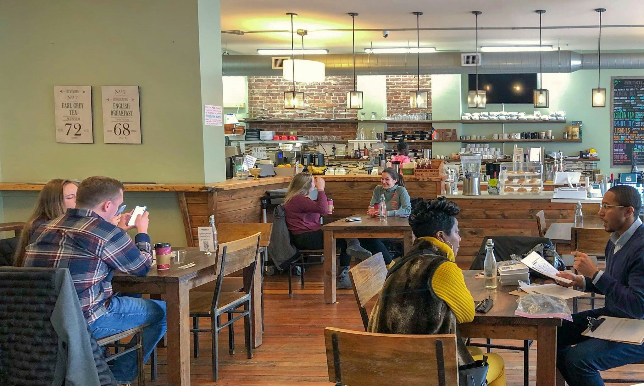 The homey interior of Arnold's Tea in Pittsburgh has light green walls, wide hardwood floors, chunky wooden tables and chairs, and wood boards lining the front of the counter, behind which are exposed brick accents, open wooden shelves lined with mugs, and a chalkboard with tea specials. Several pairs of patrons fill the tables in the forground