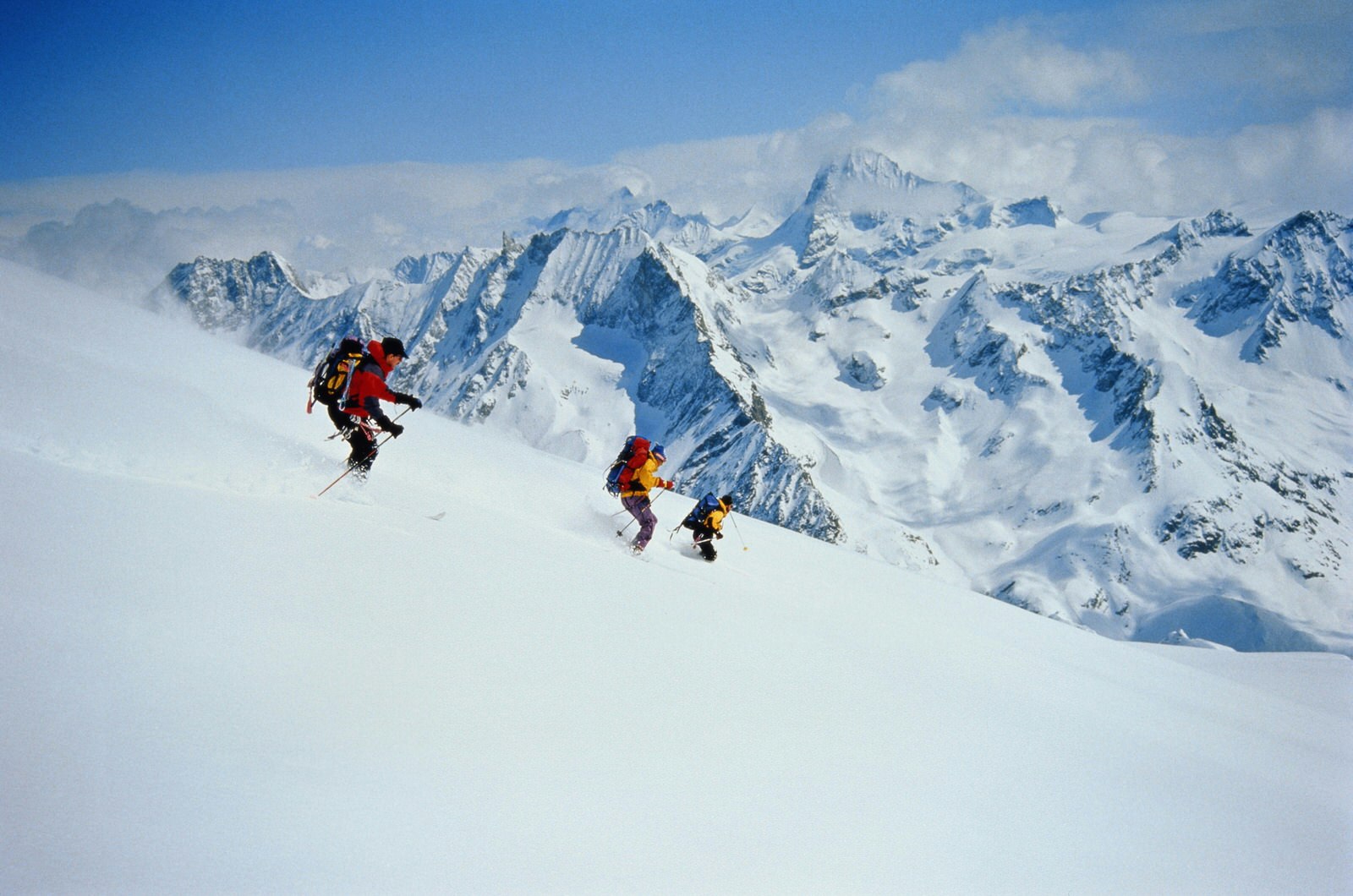 Three skiiers kitted out with backpacks ski through deep powder on a slope with mountain views in the distance
