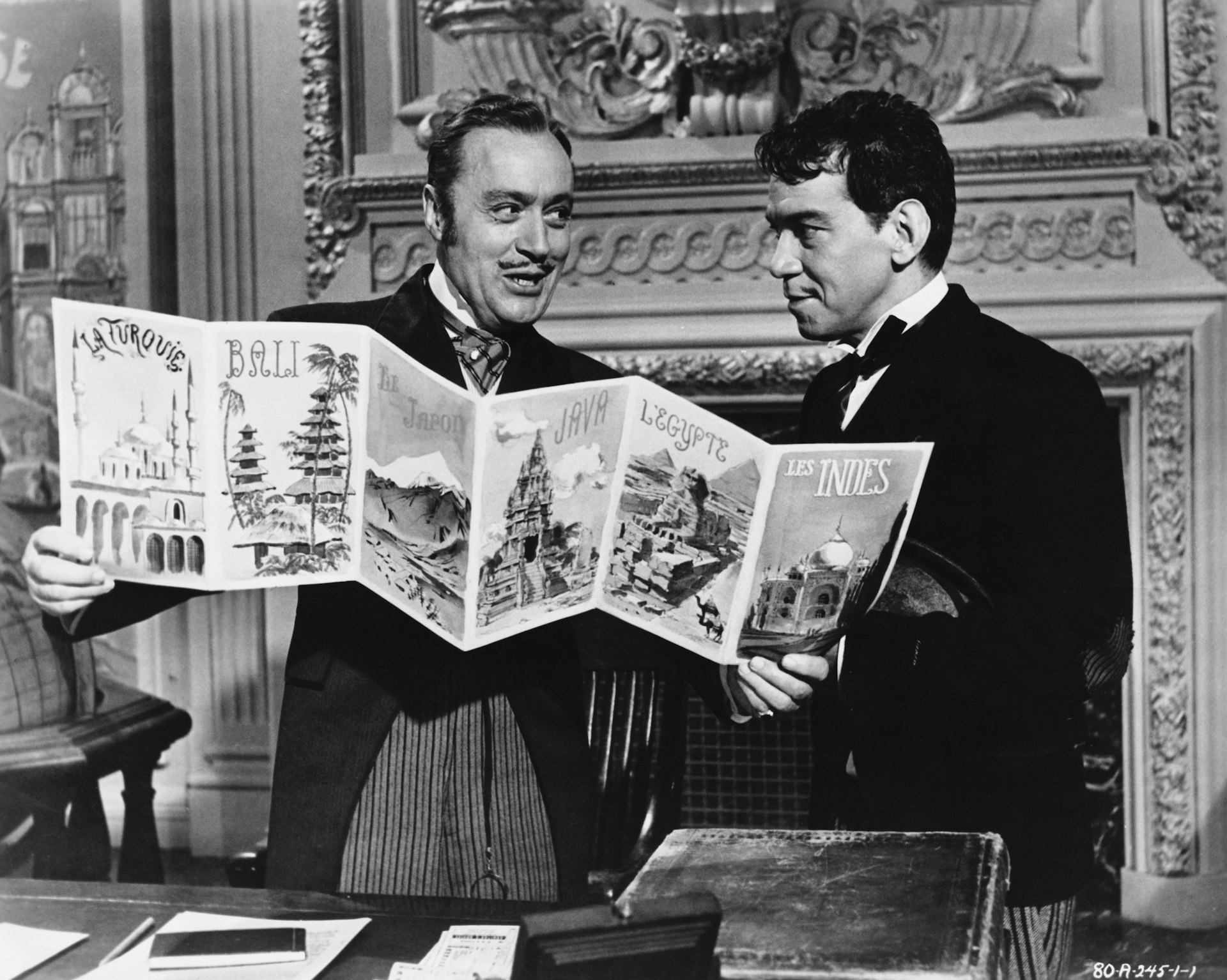 A black and white still from the 1956 film version of Around the World in 80 Days. Monsieur Gasse and Passepartout are two gentleman in black coats and ties with slick hair and are holding a brochure advertising destinations like La Turouis, Bali, Japan, Java, Egypt, and India.