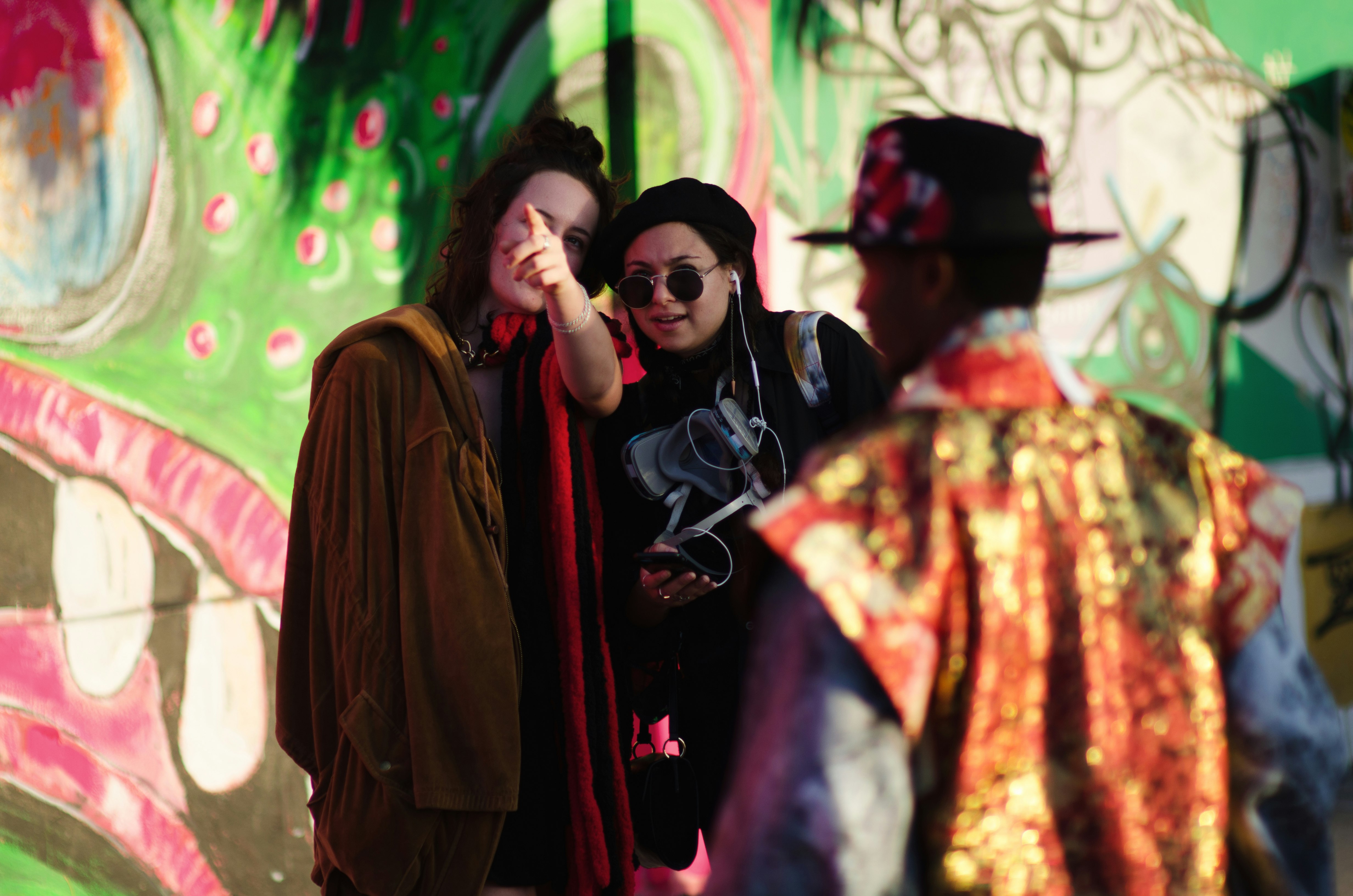 Two women are standing in front of a colourful mural at the Art Basel festival in Miami and discussing something out of the frame. A man in a bright costume is walking across the shot.