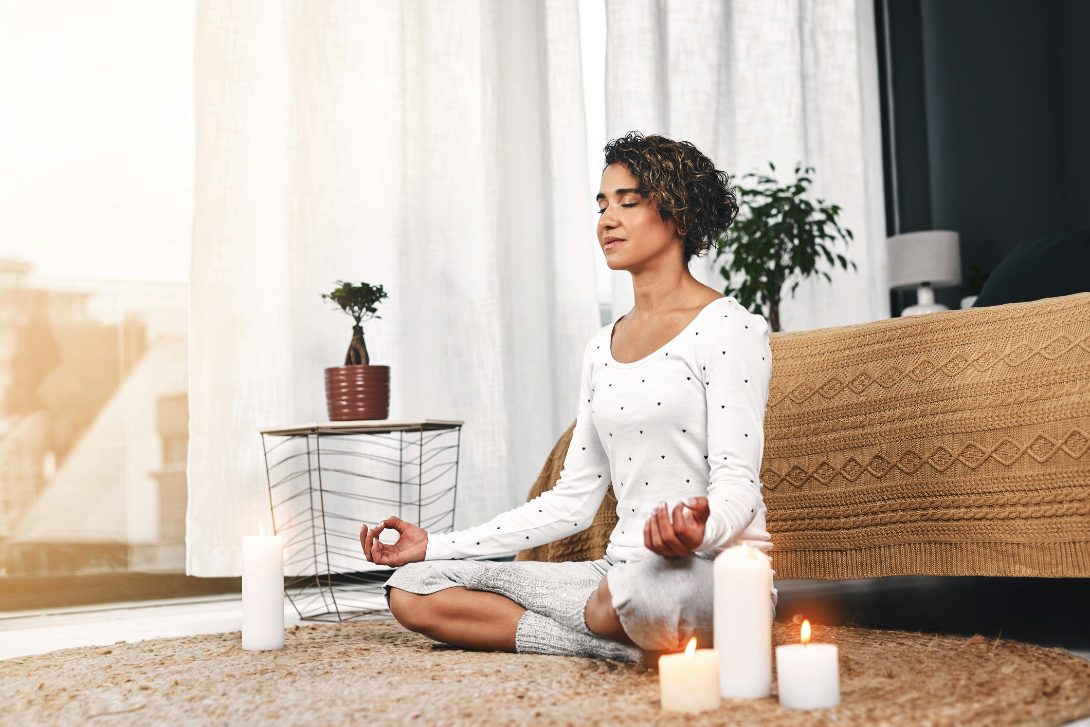 A woman with short dark hair sits on the floor of her bedroom meditating, surrounded by candles and decor in soothing neutral tones