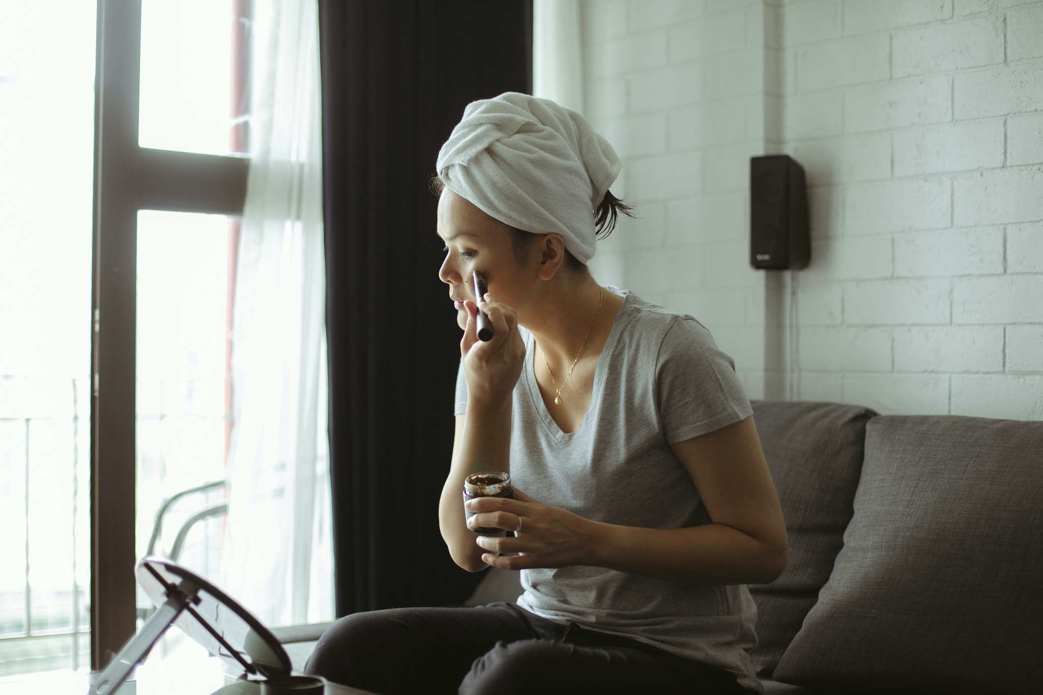 A woman sits on her living room couch and carefully applies a mud mask using a makeup mirror on the coffee table