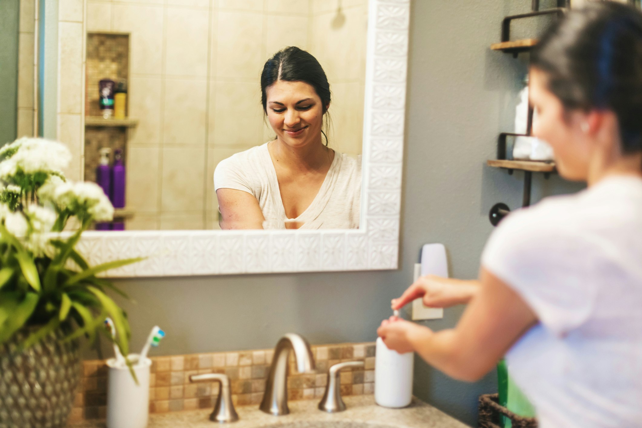 A smiling woman reaches for a product on her counter in her bathroom