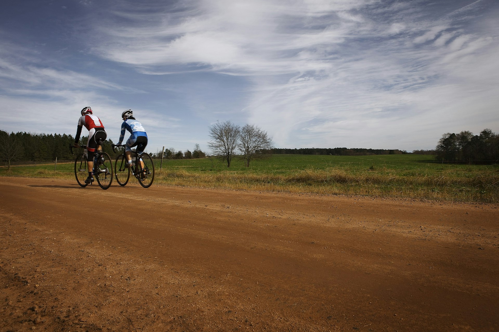 Two cyclists pedal along a red-coloured dirt road in the countryside.