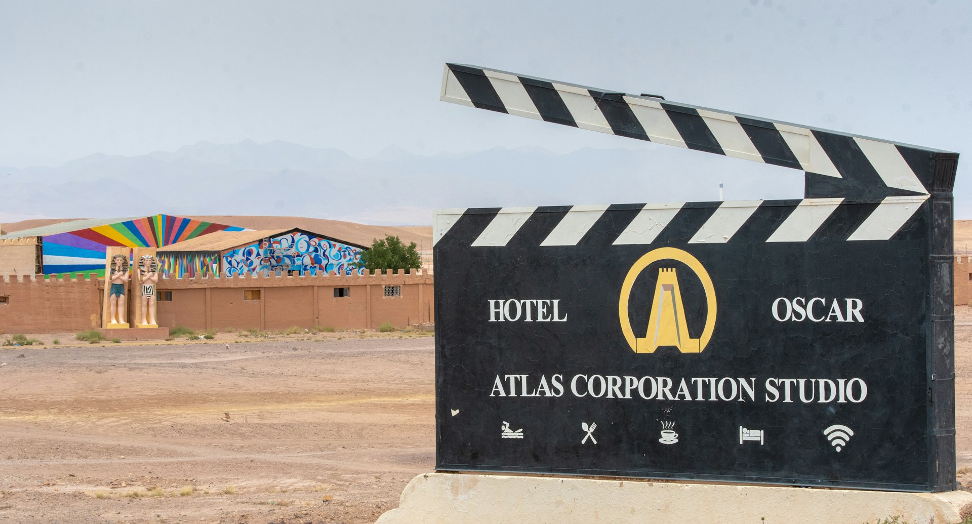 The entrance to Atlas Studios in Morocco looks like a large black clap board with a stylized illustration of a golden tower like those seen at nearby Ait Ben Haddou. Behind the sign is a crenelated wall, Egyptian style statues, and a rainbow sunburst mural  