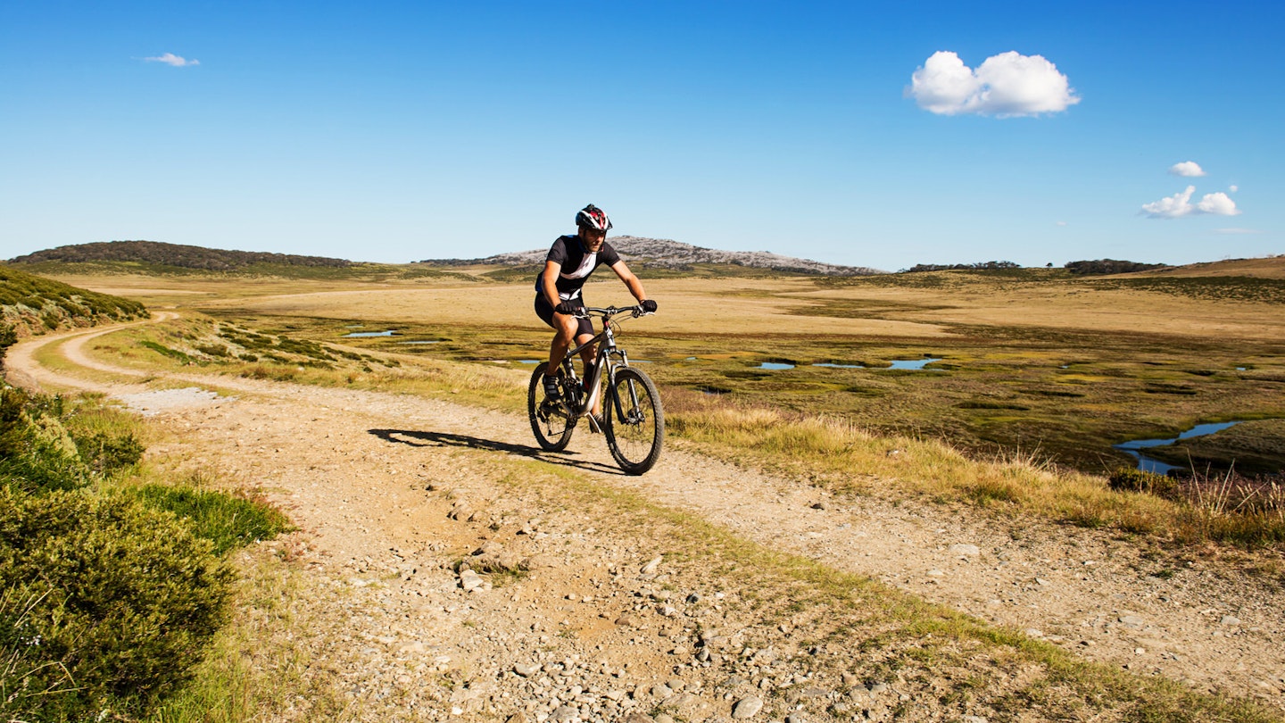 A biker is cycling along a rough track in the Australian countryside on a sunny day.