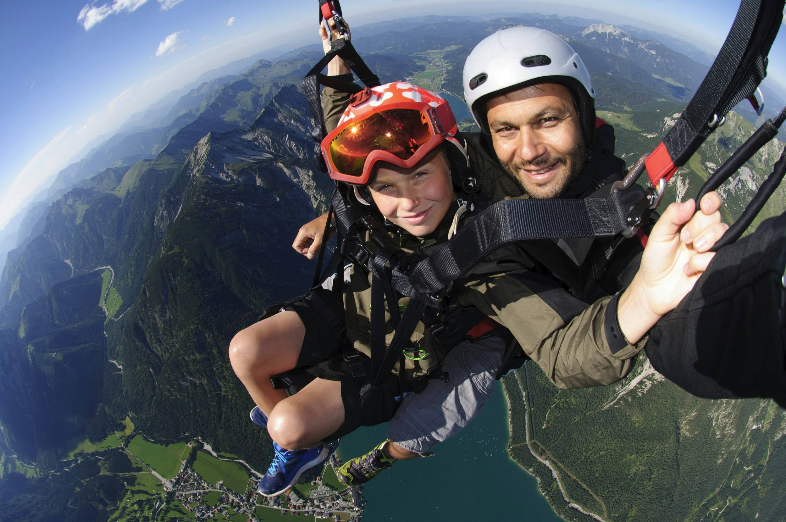 A paragliding pilot together with his youngest son, high up with a tandem paraglider above lake Achensee in Tirol, Austria. The pilot and the child are looking into the picture with a big smile. In the background mountain landscape in summer.