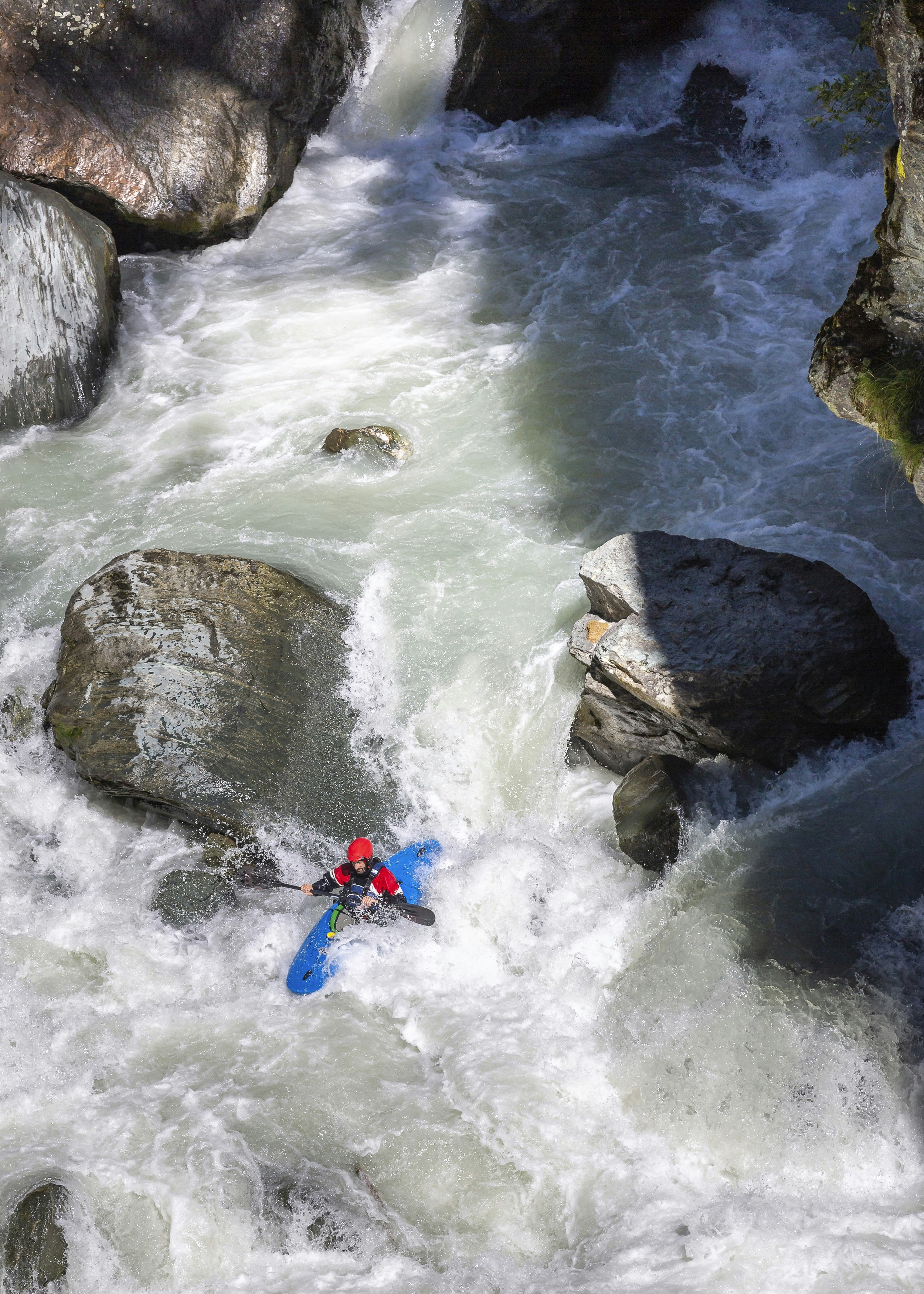 Vertical shot of a male athlete running whitewater rapids in Austria; the water is frothing as it cuts between a series of large boulders in the river.