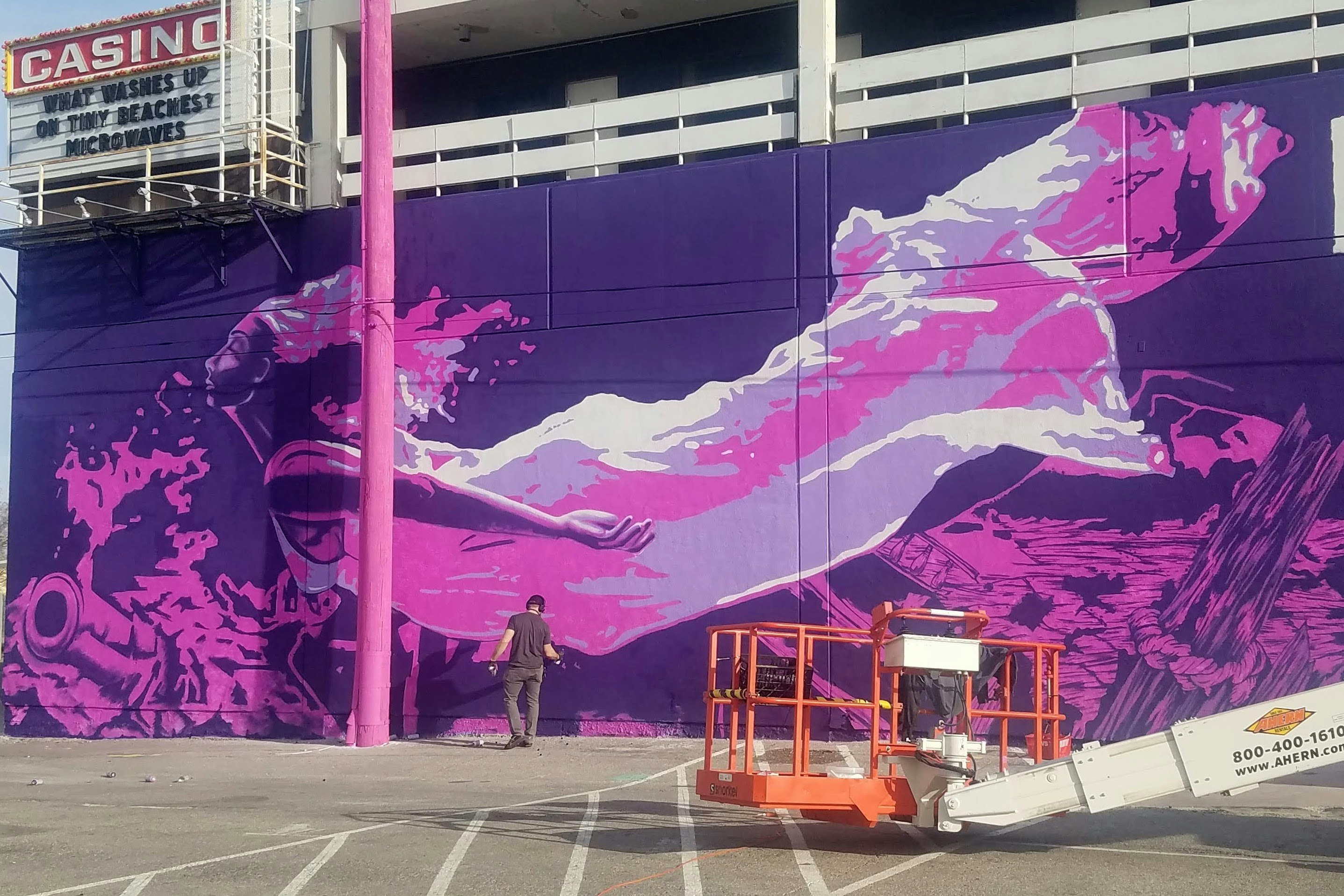Artist Aware adding the final touches to a huge purple and pink mural; it depicts what appears to be a woman swimming in a floaty gown.