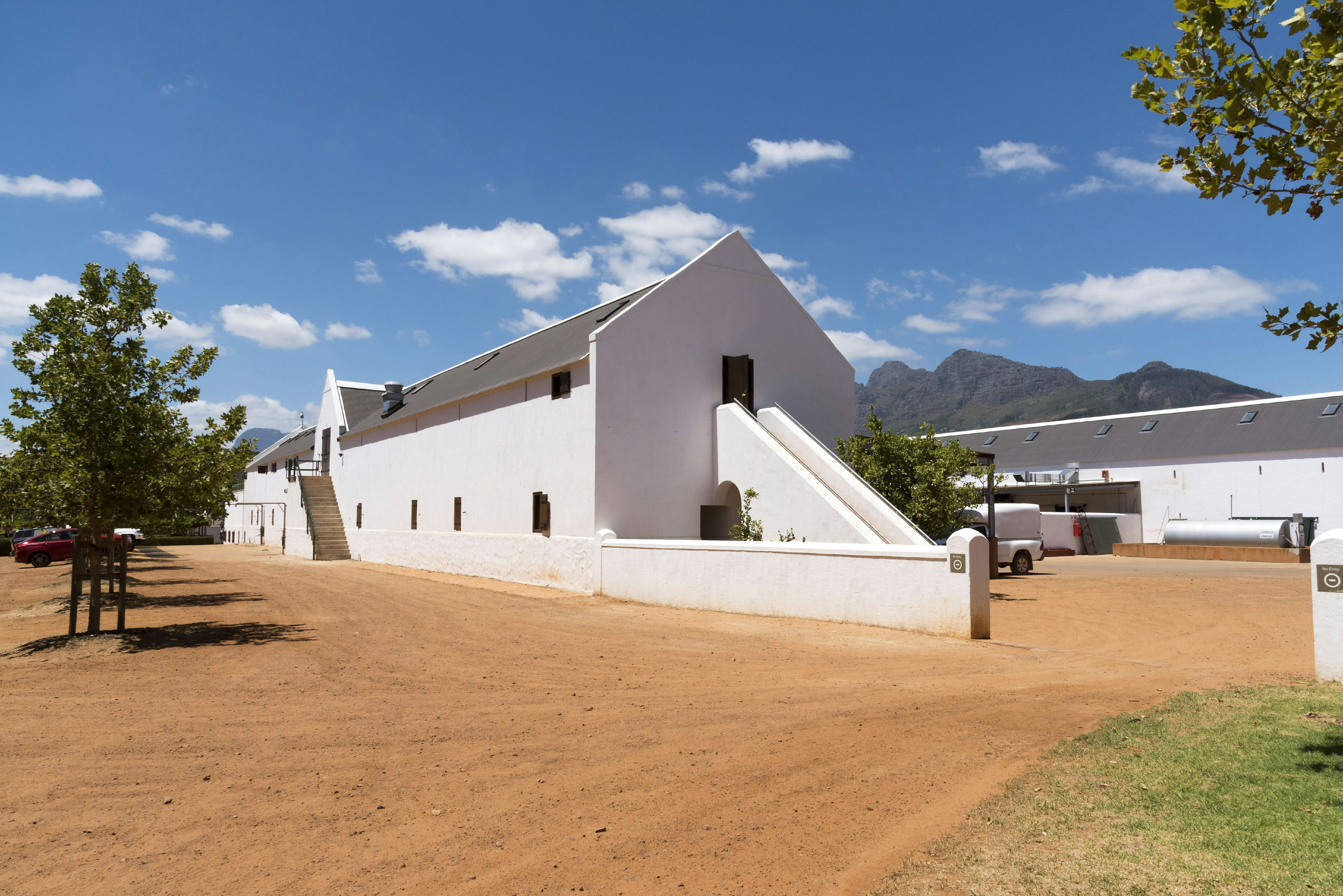 A white adobe building at Babylonstoren situated on a red dirt road with a small tree in the foreground and green hills in the background