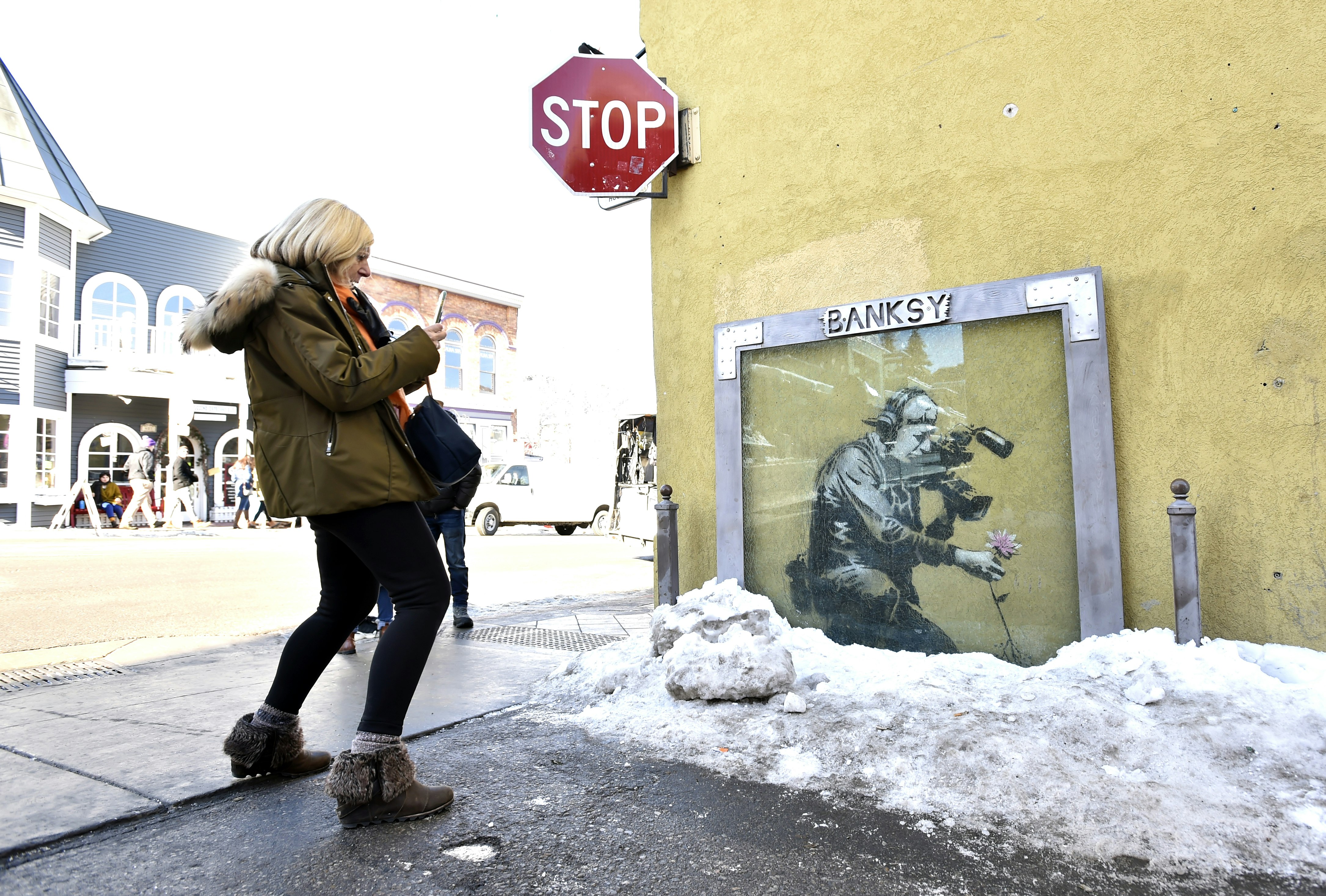 A woman in wedge ankle boots and a olive parka with blond hair leans back and bends her knees as she snaps a photo of a Banksy mural in Park City