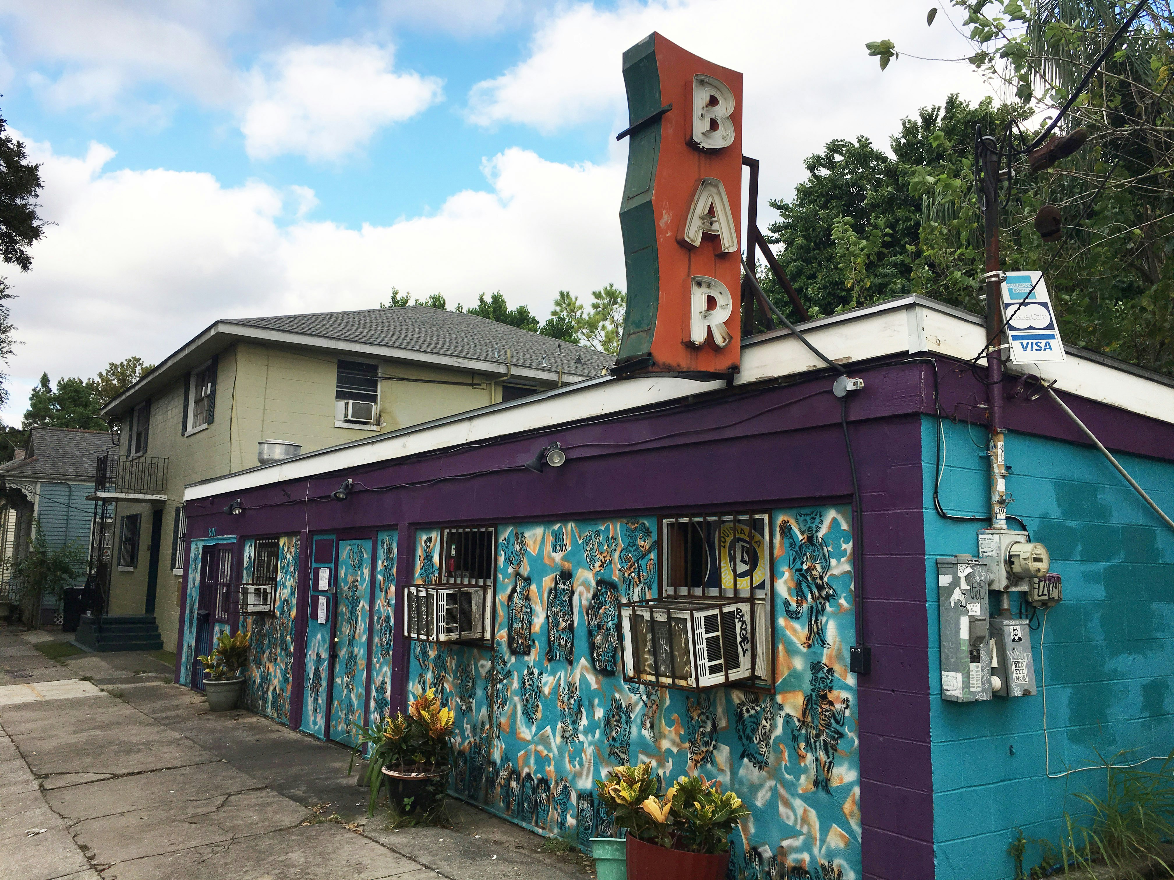 Exterior shot of Bar Redux with its bright blue bricks and purple boarder. There are small black figures painted on the front part of the building and a large "Bar" neon sign on the roof. 
