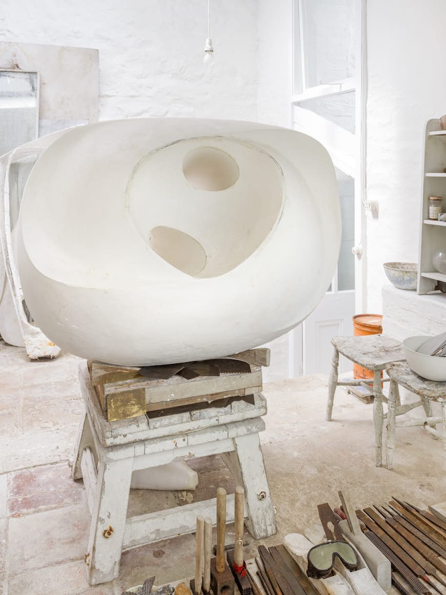 Интерьер блестящей белой студии в музее Барбары Хепворт;  на деревянном табурете сидит большая белая выпуклая скульптура.