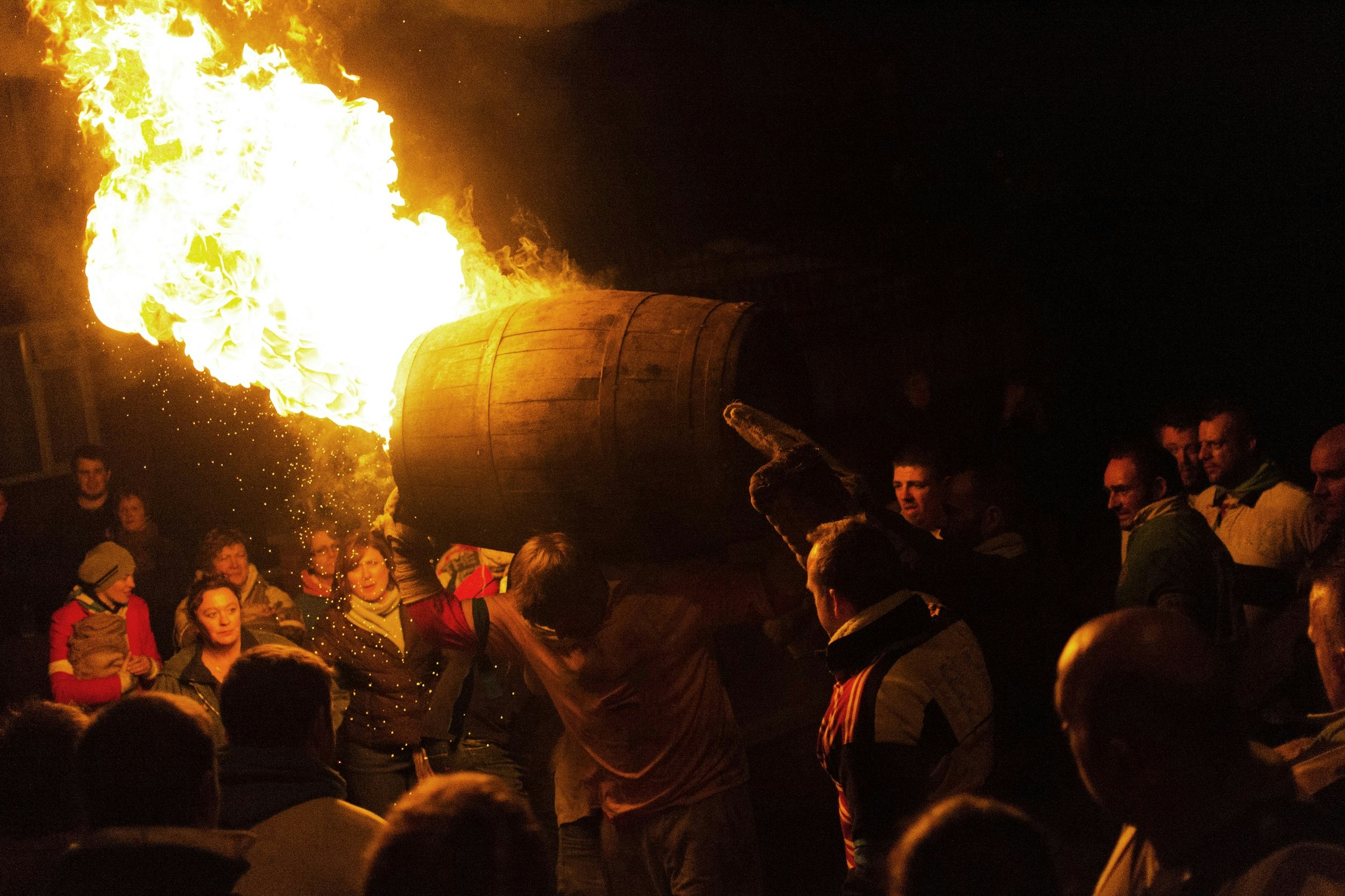A man has a burning barrel on his shoulders as he walks through a crowd of people