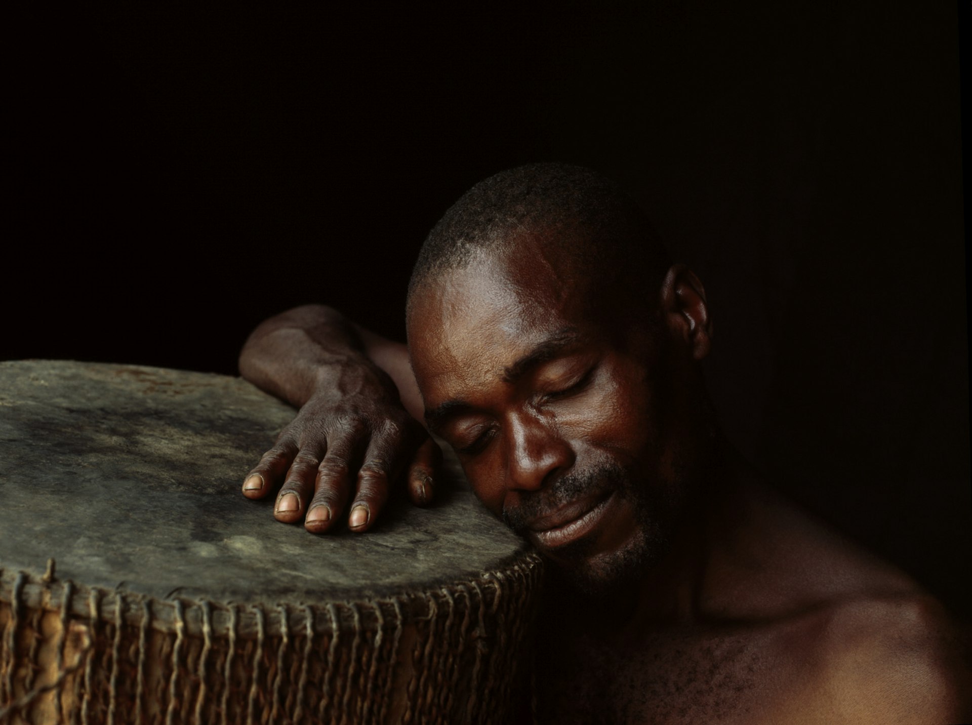 A Batwa man sleeps with his head on his drum; he has a peaceful smile on his face