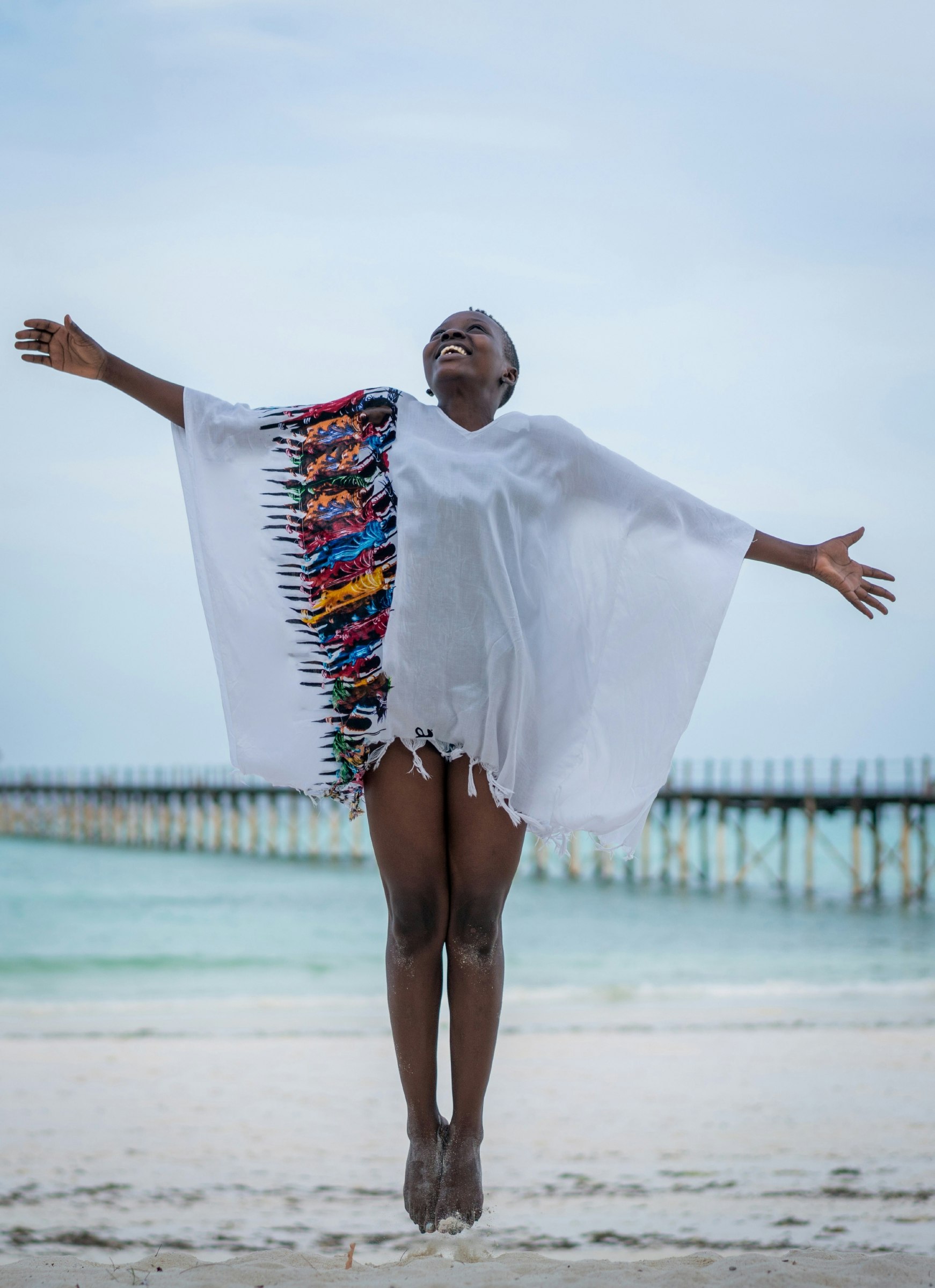 Young woman jumping on a white sandy beach with a pier in the background..jpg