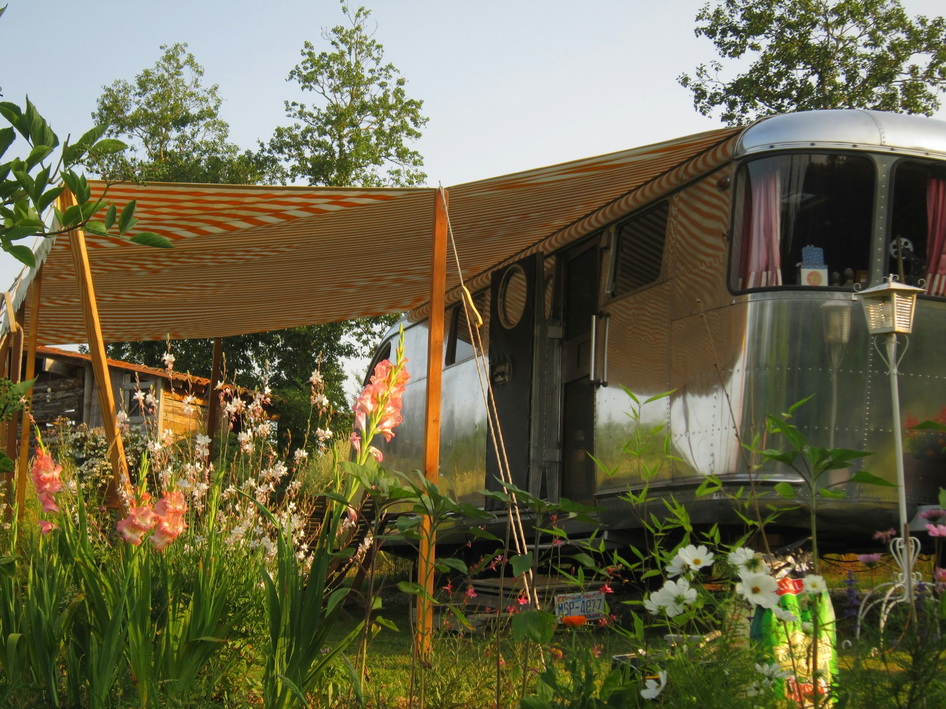 A silver Airstream camper with a white and orange striped awning extended from the left side sits in a garden patch of wildflowers and coral-colored gladiolas. In the background a wooden bath house adds to the rustic charm. 