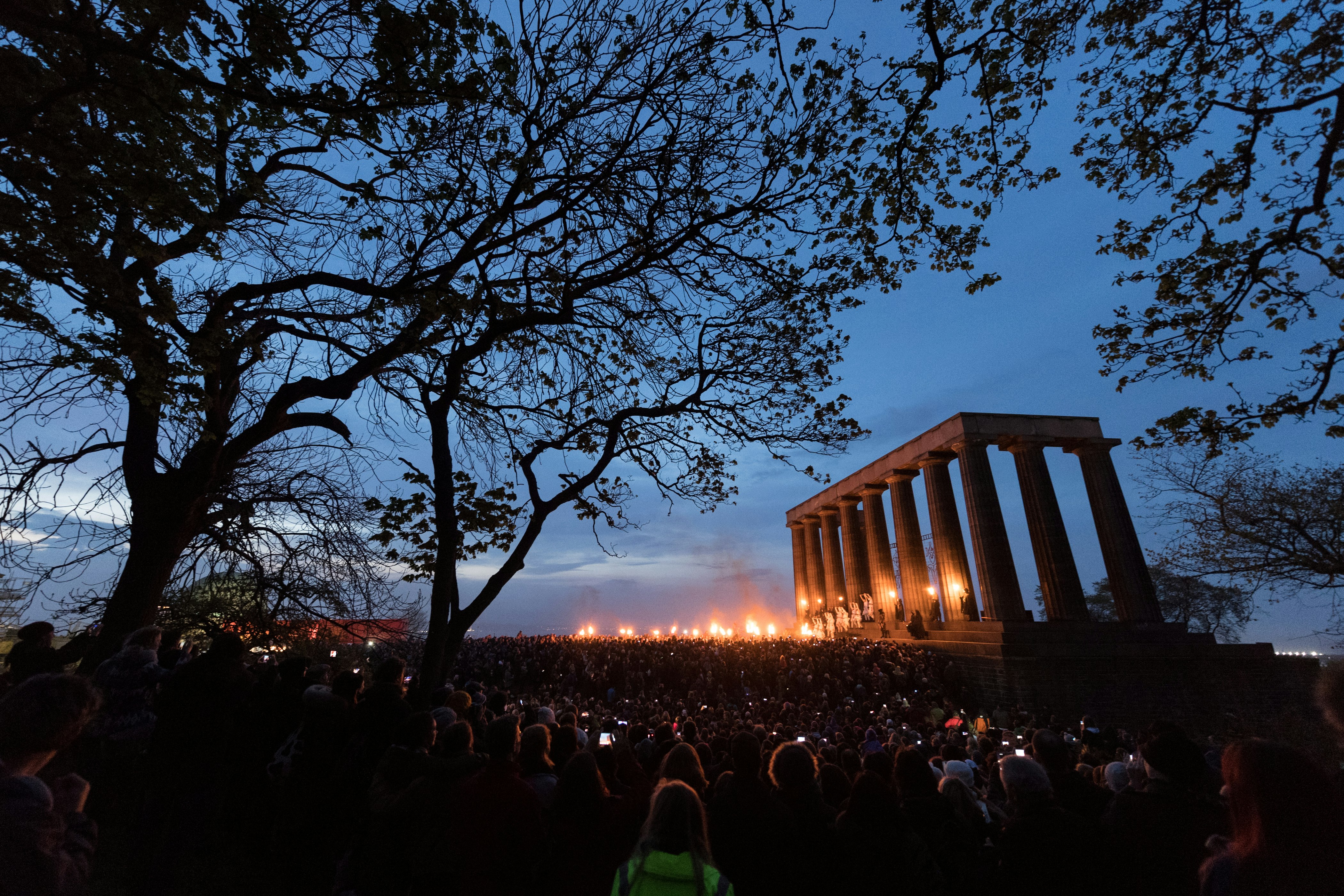 The partially completed columns of the Parthenon replica on Carlton Hill is lit by bonfires and torches that are faintly illuminating a vast crowd, some holding glowing cellphone screens against the light blue dusk