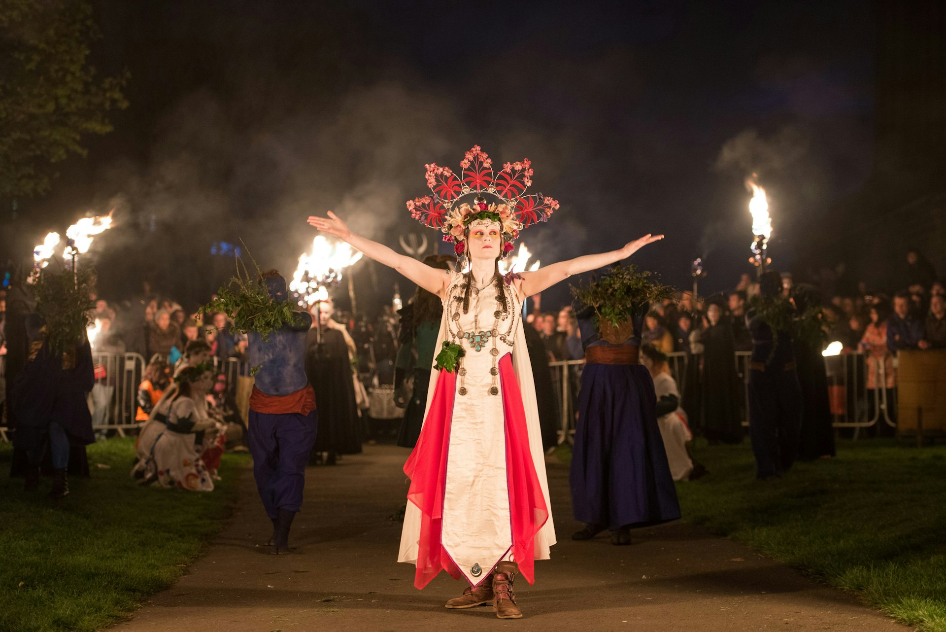 The May Queen raises her arms in greeting as she processes through the crowds at Beltane Fire Festival. She has a large headdress in the shape of red flower petals, a white dress, and red fabric hanging down in front. Behnd her large flames burn against the black night sky and people stand watching