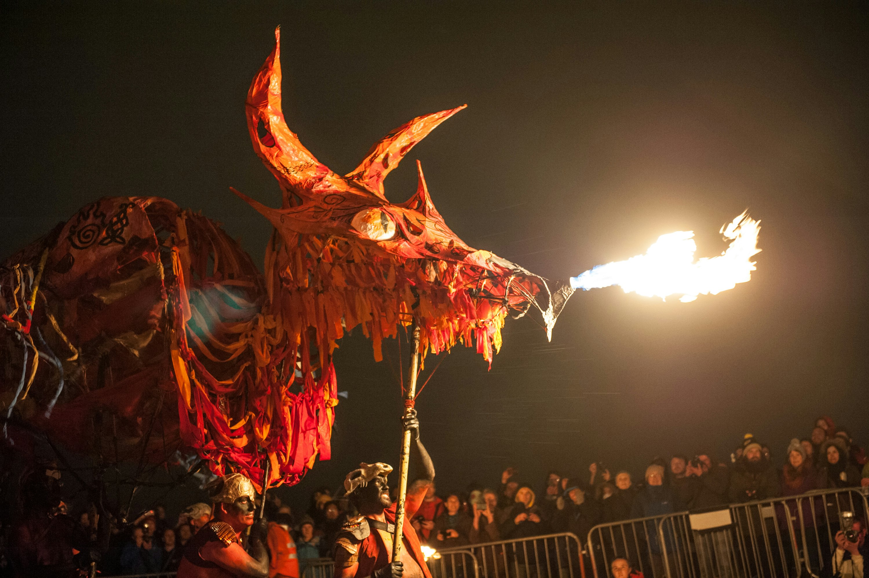 A fire-breathing dragon puppet made of cloth pieces in orange, pink, and read draped over a wooden frame is held aloft by performers in body paint and masks. In the background are metal security barriers and a crowd of observers.