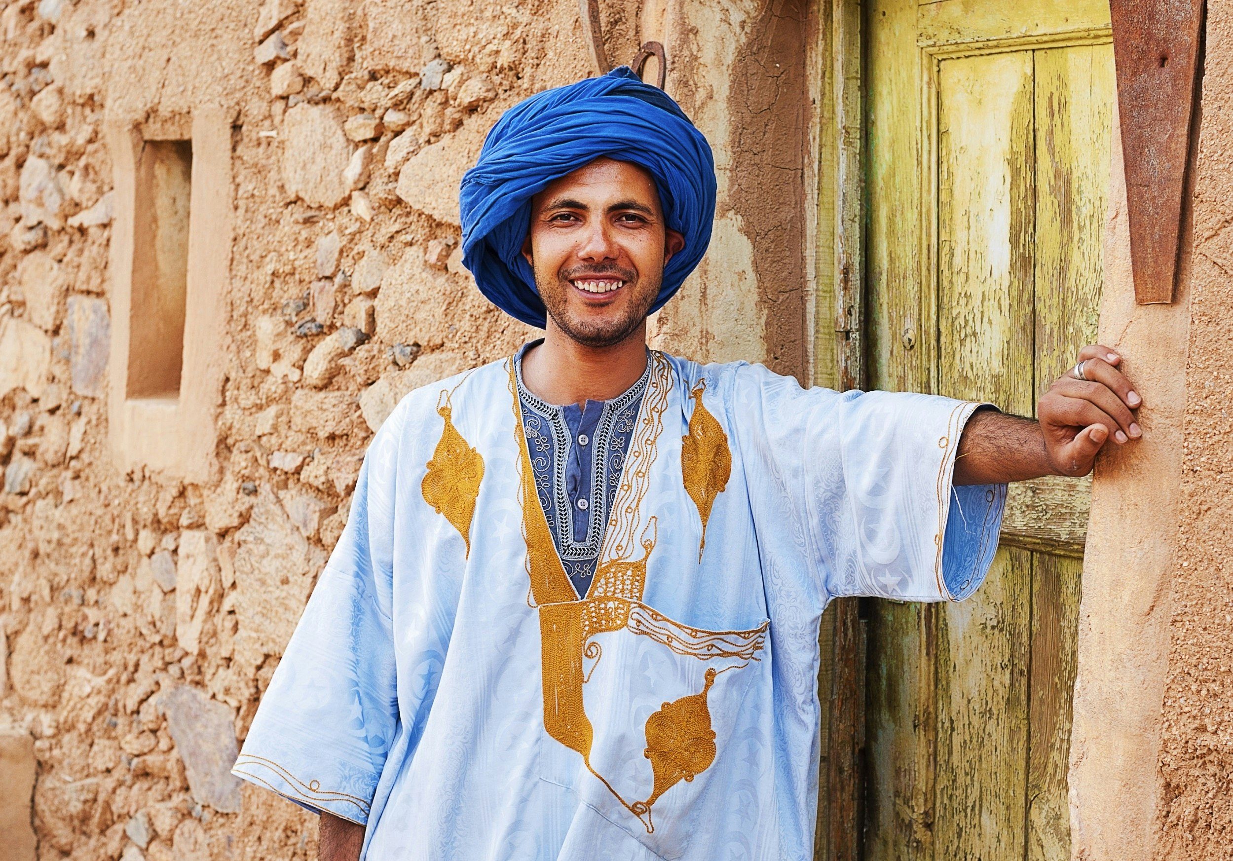 A Berber man, with indigo head scarf and light blue and gold robe stands smiling in front of a pinkish stone building.
