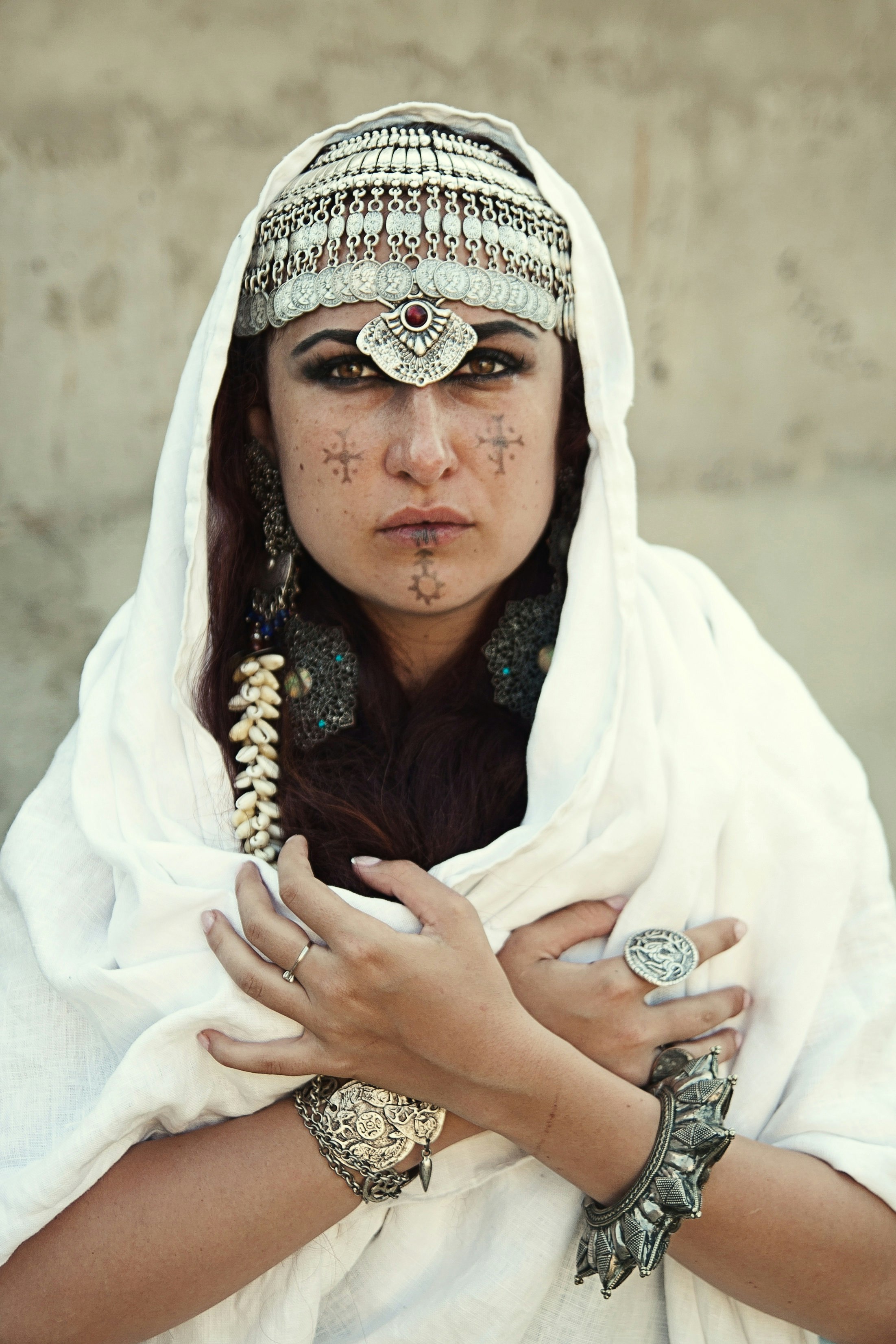 A Berber woman stares into the camera, her cheeks and chin each donning a traditional symbol; she wears a decorative headpiece and large sliver bracelets. Her arms are crossed over her chest and a white shawl covers the top of her head, shoulders and torso.