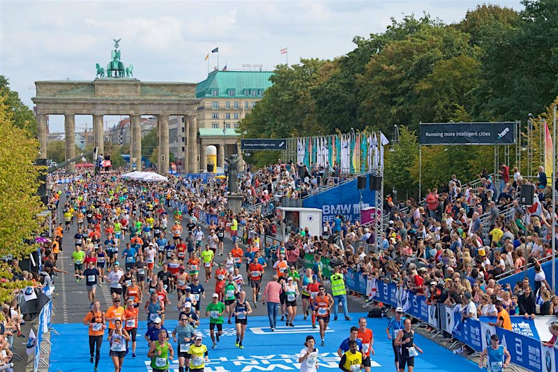 Thousands of runners stream through the Brandenburg Gate and run towards the camera down a tree-lined boulevard.