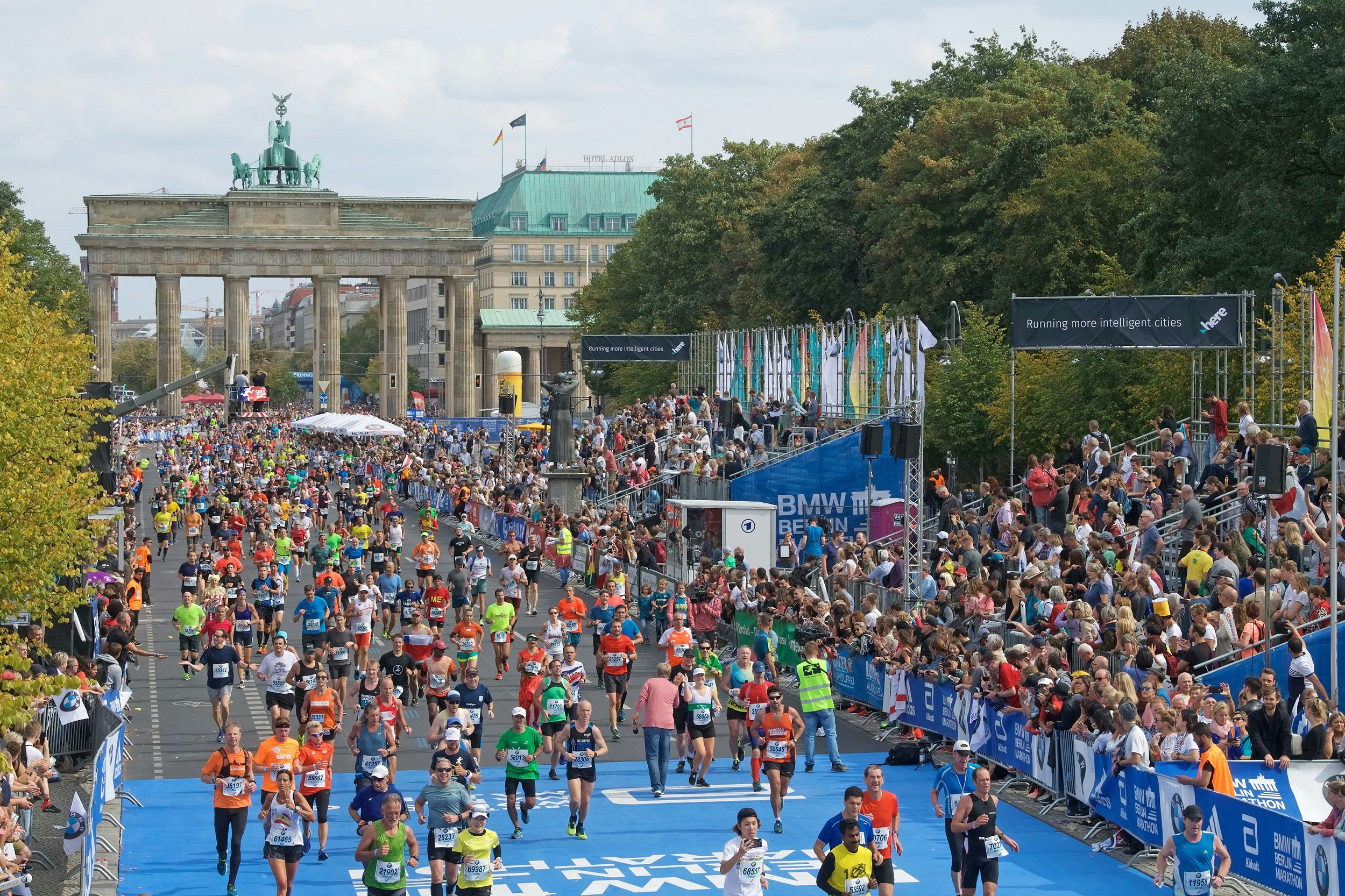 Thousands of runners stream through the Brandenburg Gate and run towards the camera down a tree-lined boulevard.