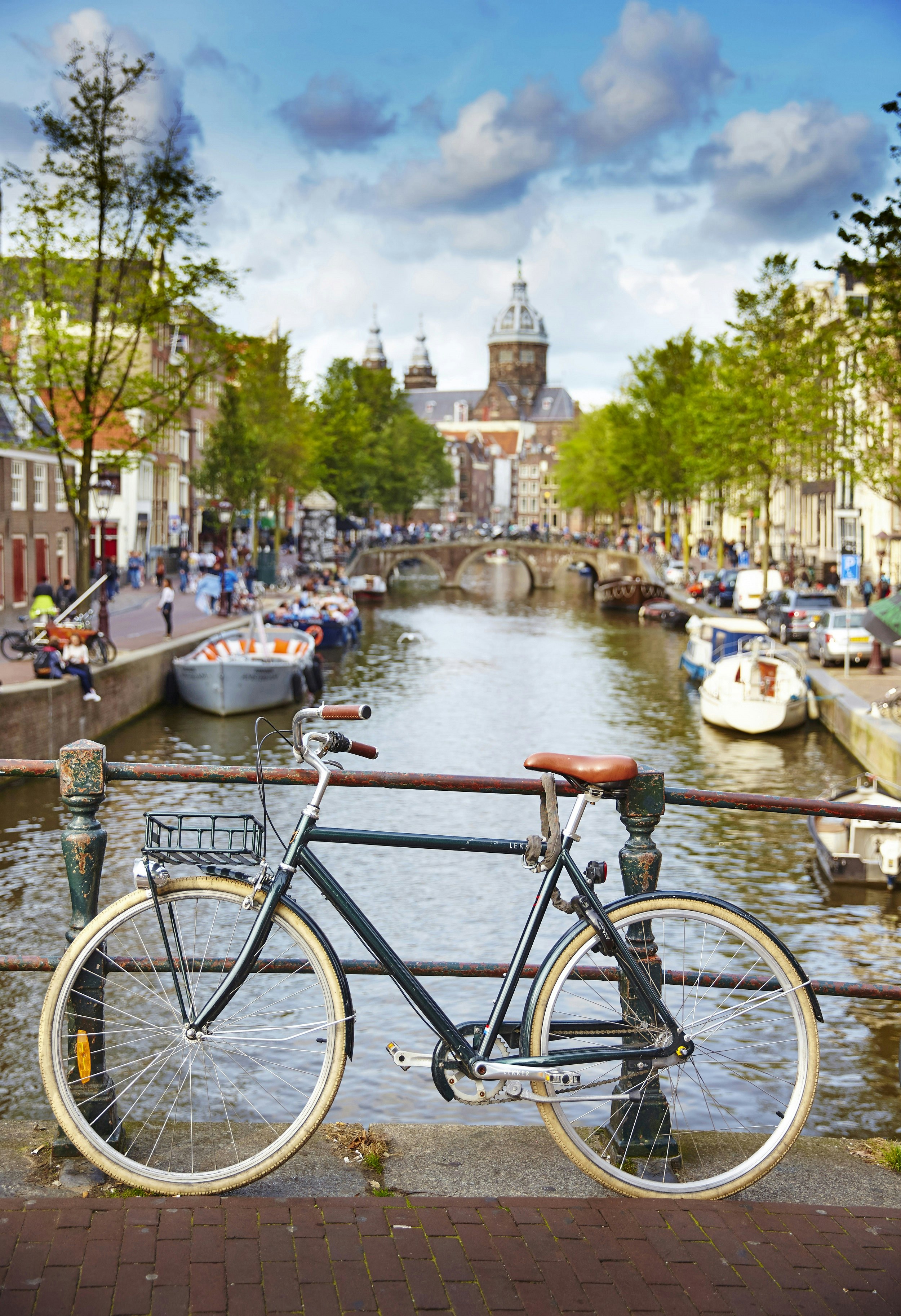 A traditional bicycle is parked against a hand rail on a bridge over one of Amsterdam's canals; boats line the canal and a large church rises from its end in the distance.