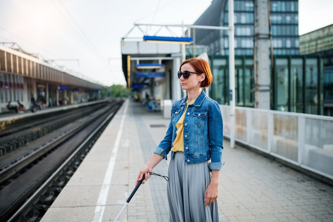 portrait of a blind woman with a white cane standing outside, waiting for public transport in summer.