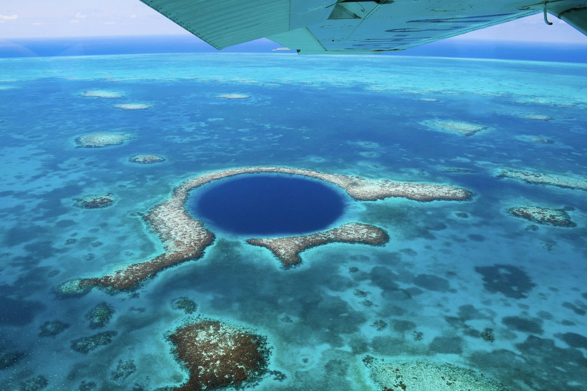 The image looks down the underside of a light aircraft's wing to the deep blue circular hole in the Light House Reef; the surrounding waters are turquoise in colour.