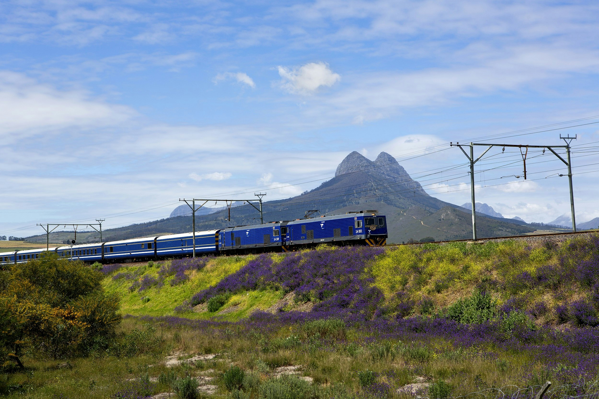 A train with blue carriages passes through stunning grassy landscapes, with high peaks in the background