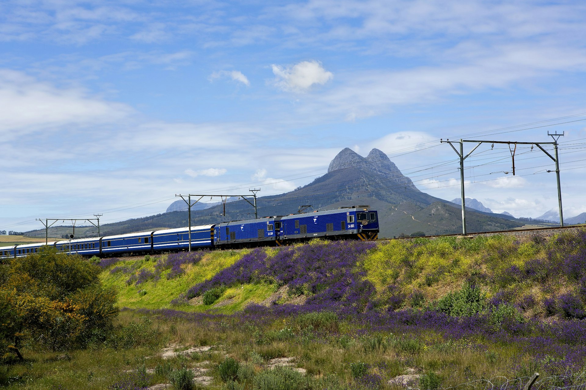 A train with blue carriages passes through stunning grassy landscapes, with high peaks in the background