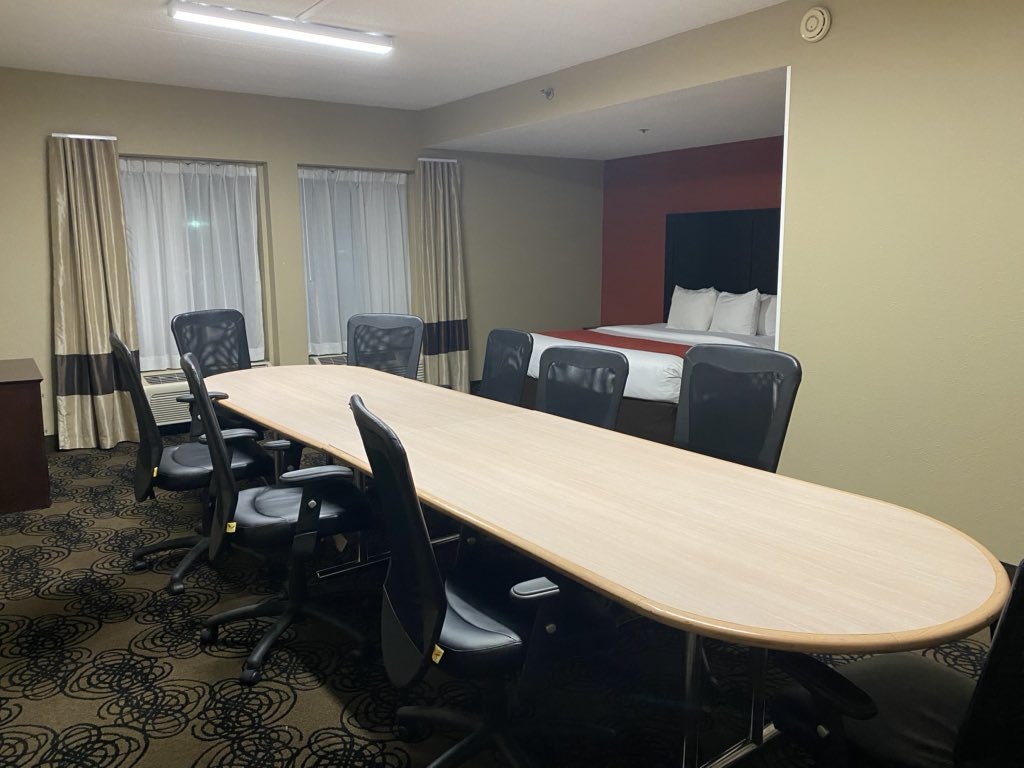 A beige-decorated room has an enormous boardroom table surrounded by office chairs in the middle of it; there is also a kingsize bed set into a recess.