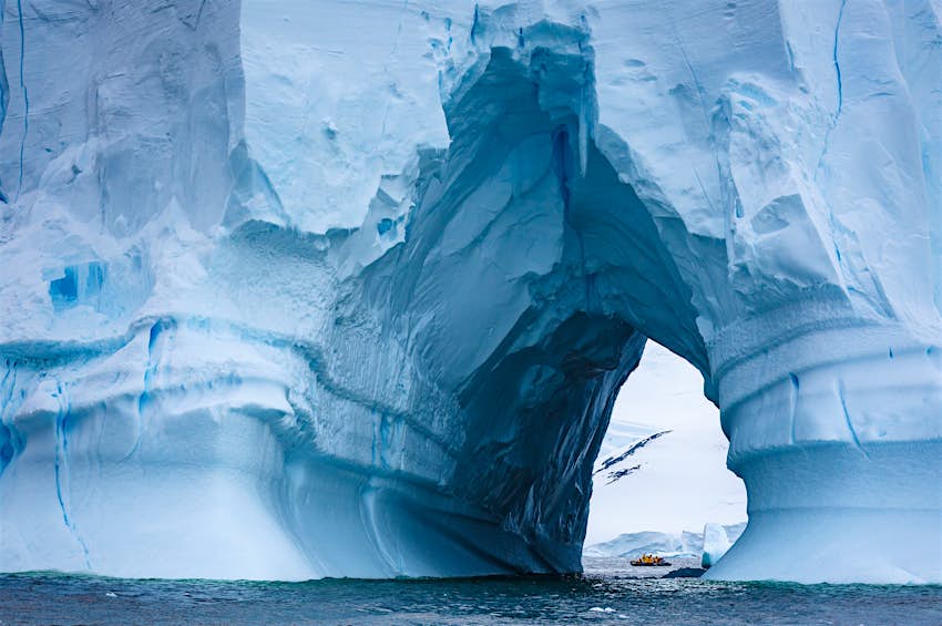 A mammoth iceberg fills the entire frame of the shot, with a natural tunnel through it revealing a zodiac boat with tourists on the other side.