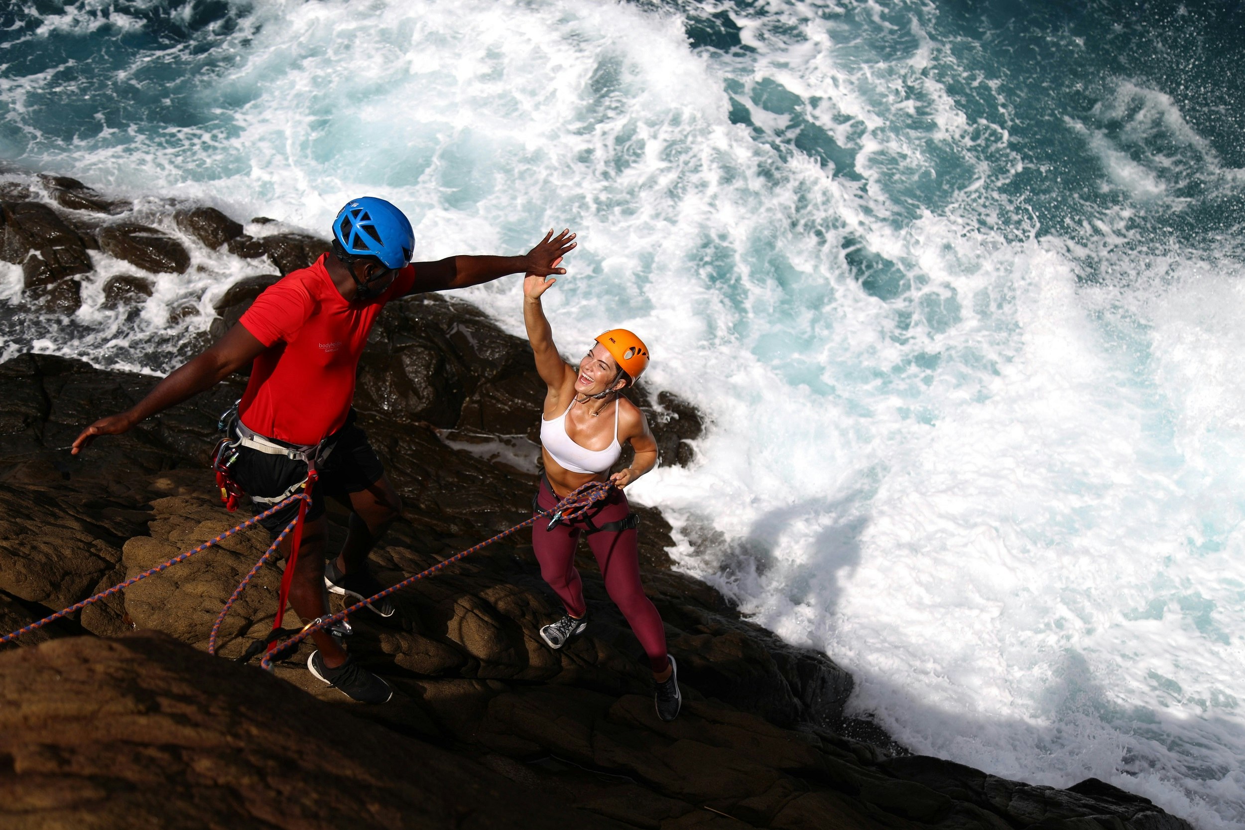 A woman wearing a climbing harness stands on a near vertical rock wall and high-fives her instructor above; below are crashing waves.